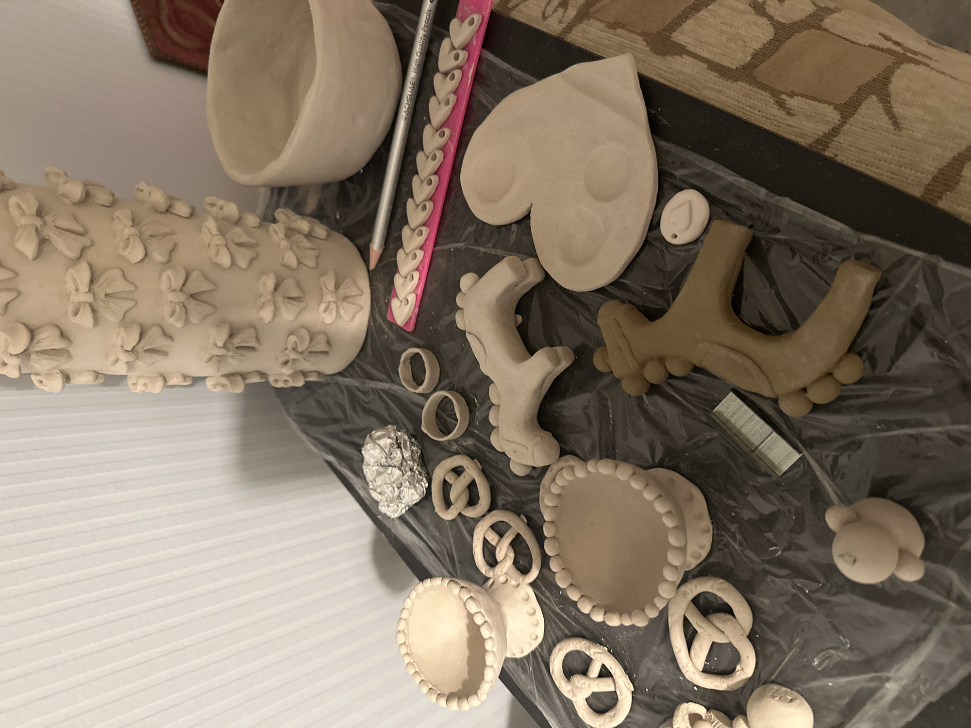 The image shows a close-up, overhead view of various unbaked clay creations and craft supplies laid out on a dark, plastic-covered surface. The objects are primarily light beige in color. Prominently featured is a tall, cylindrical vase decorated with numerous small, bow-shaped embellishments. To the right of the vase is a simple, unadorned bowl.
Scattered across the surface are several clay pretzels, two small clay rings, and two stylized clay horse figures. One horse is a lighter, more finished shade of beige, while the other is a darker, possibly slightly drier clay. A large, flat heart-shaped clay piece with two circular indentations is positioned near the horses. There is also a small, oval clay charm with a hole.
In the foreground, a ceramic stand with a beaded rim holds a small bowl. Another similar stand with a beaded rim is partially visible to the left. Further to the left, a small, spherical clay figure resembling a bear or similar creature is visible. A crumpled piece of aluminum foil is located near the top center of the arrangement.
A pink ruler with small heart cutouts is placed diagonally across the middle of the scene, with a silver pencil resting on top of it. A small stack of metal staples is situated near the bottom center. The background consists of a textured, light-colored surface, possibly a wall with vertical blinds. The overall setting appears to be a craft area or workspace. There is no indication of the specific location in Nevinnomyssk, Russia, as the image lacks any geographical markers.