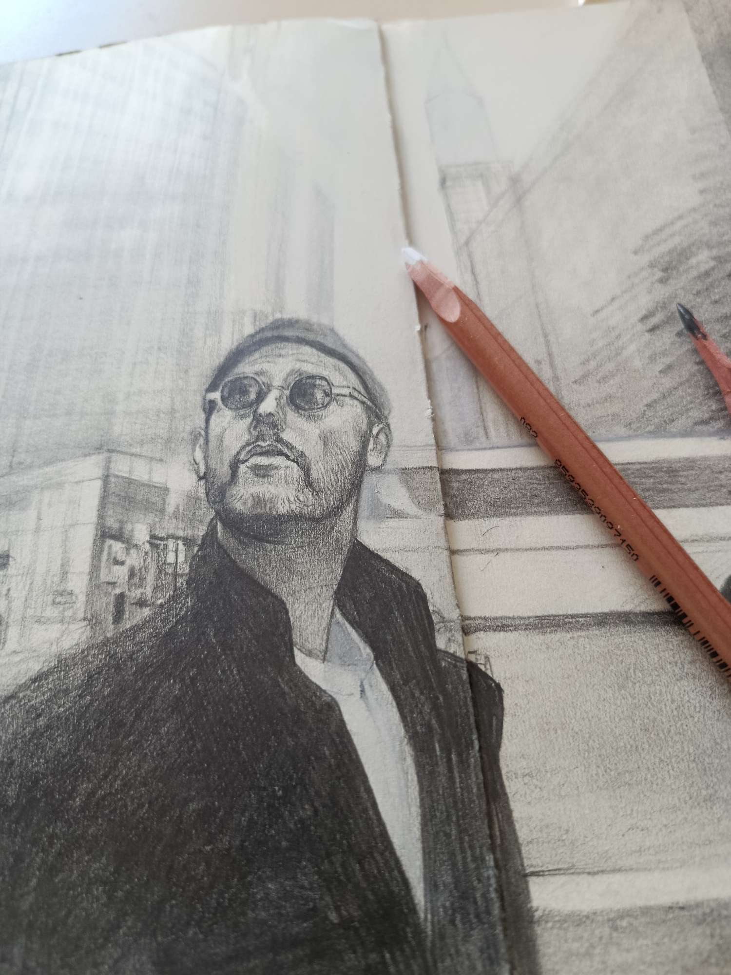 A detailed pencil drawing of a man, resembling actor Jean Reno as his character Leon from "Léon: The Professional," is the central subject of the image. The man is depicted from the chest up, looking upward and slightly to the left, with a serious, contemplative expression. He wears a dark beanie, distinctive round sunglasses, and has a full beard and mustache. His attire includes a dark, heavily shaded jacket or coat over a lighter-toned shirt.
The drawing, rendered with realistic shading and fine pencil strokes, occupies the left side of what appears to be an open sketchbook or two pieces of paper joined at the center. The background features a subtly sketched cityscape with faint lines suggesting tall buildings and urban structures, providing a sense of depth.
Two pencils are visible in the foreground, resting on the right side of the artwork. The most prominent one is a light-brown wooden pencil with a sharply pointed white tip, indicating a soft white lead. Printed text "002" and "8593539007150" is clearly legible on its barrel, along with small barcode-like markings. A second pencil, with a darker tip, is partially visible further to the right.
The paper has a visible texture, enhanced by the natural lighting which illuminates the scene from the upper left. The overall impression is that of an in-progress or recently completed artistic rendition, captured intimately with the artist's tools. The setting appears to be indoors, likely where the art is being created or displayed.