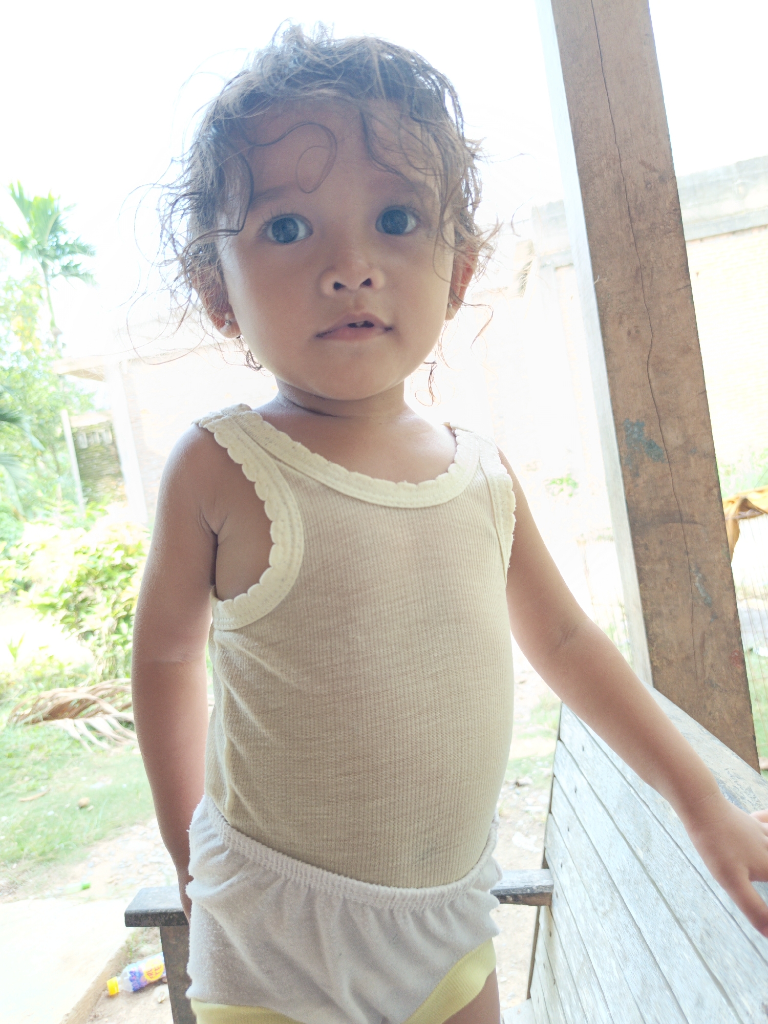 In Serbajamantunong, Indonesia, a young child with curly brown hair, wearing a cream-colored tank top and white shorts with a yellow trim, stands facing the camera. The child's eyes are open and looking forward. The background features lush green vegetation and a wooden structure, suggesting an outdoor setting. The lighting is bright, indicating daylight. The child appears to be standing on or near a wooden bench. To the left of the bench, a plastic bottle with a yellow cap is visible on the ground. The overall scene is informal and casual.