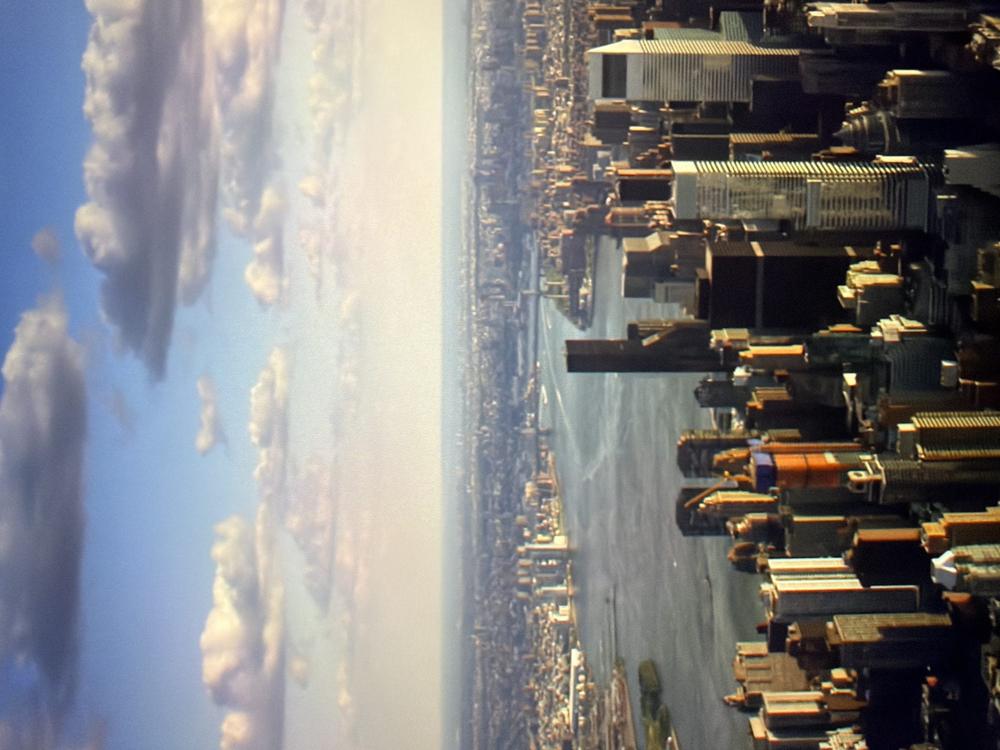 This is an aerial, wide-angle photograph of a dense cityscape with a body of water in the foreground. The sky above is a bright blue with scattered white and gray clouds. The cityscape features numerous buildings of varying heights and architectural styles, with many appearing to be skyscrapers. Some buildings are illuminated by sunlight, casting shadows, while others are in shadow. The water in the foreground appears to be a river or a bay, with a few small boats visible. The distant horizon shows more urban sprawl. The image captures a sprawling metropolitan area from a high vantage point, suggesting an urban environment with significant development. The image does not depict the city of Kyiv, Ukraine.