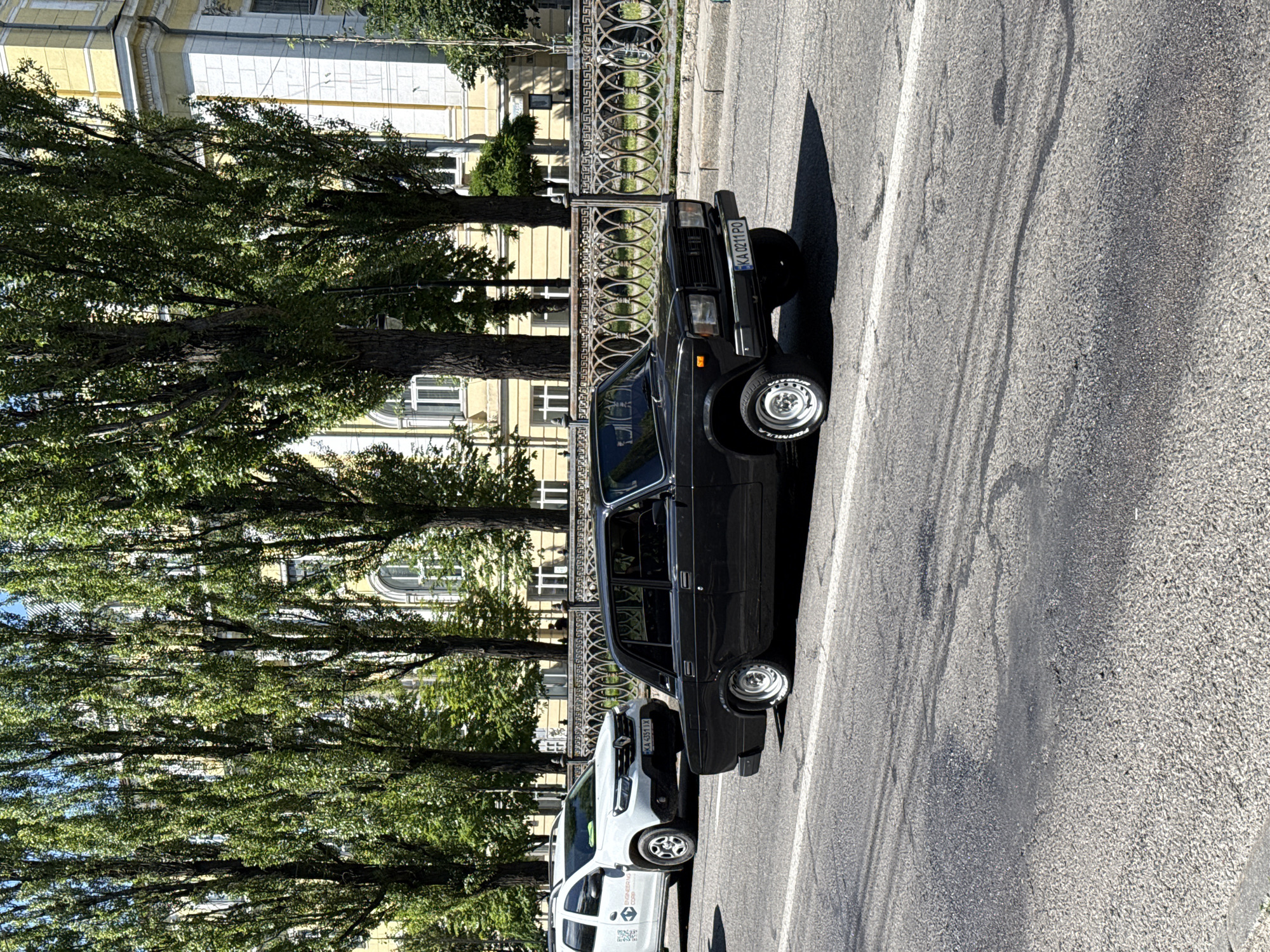 A black sedan with a license plate reading "KA 0211 PO" is parked on a city street in Kyiv, Ukraine. The car appears to be a Lada. Behind the car, a white SUV is visible, with a license plate that reads "KA 4351 IX". In the background, tall trees line a street, with a building visible behind the trees. The building has yellow walls and white trim. A decorative metal fence runs along the sidewalk. The street is paved and appears to be in good condition. The sky is clear and blue, suggesting a sunny day.