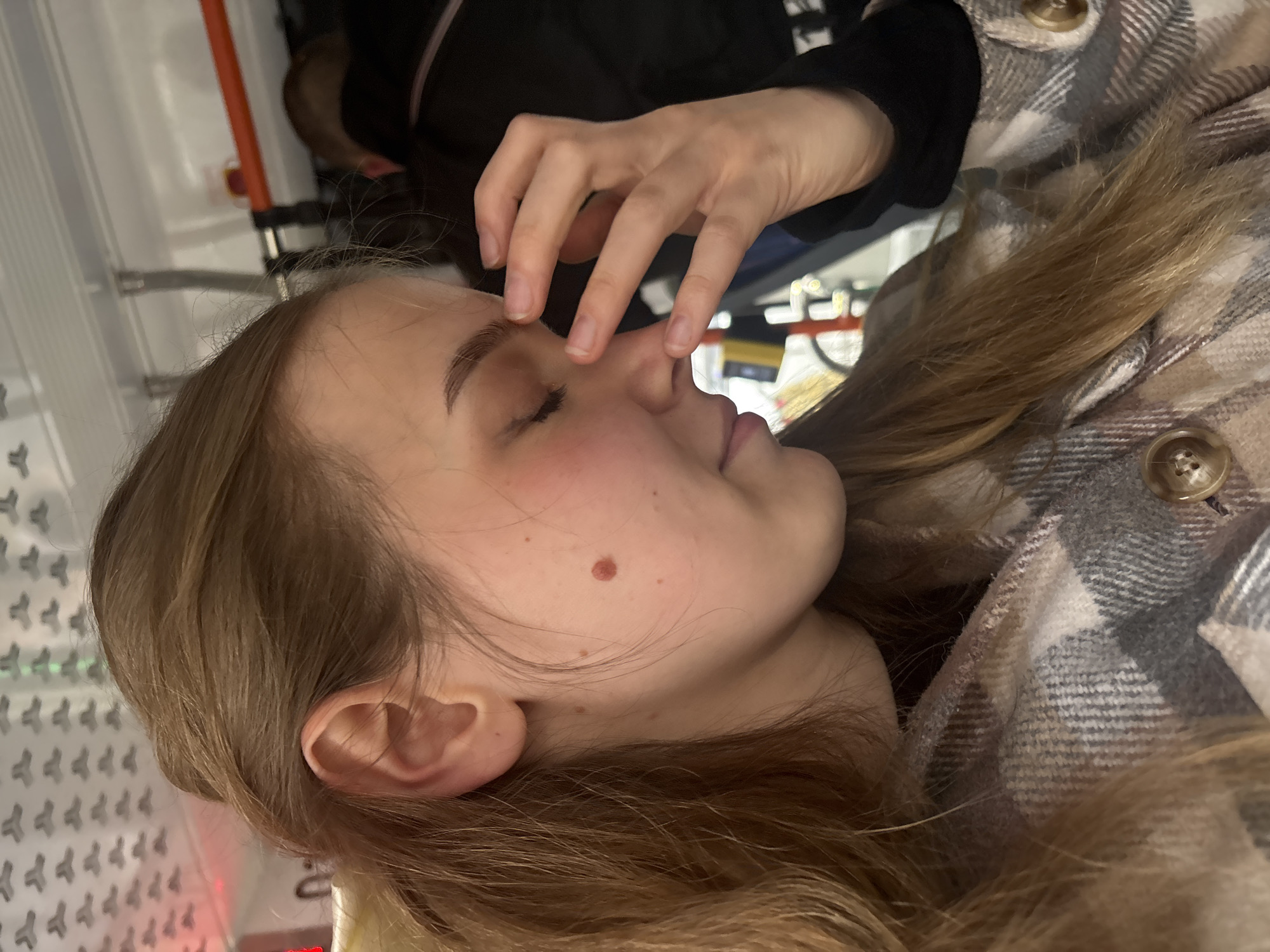 A close-up, indoor shot depicts a young, light-skinned female with medium-length brown hair. She is wearing a patterned flannel jacket with visible buttons. Her eyes are closed, and her hand is positioned near her face, with fingers touching her nose. A mole is visible on her cheek. The location is Unknown, Russia. The background shows a metallic structure and suggests a medical or institutional setting.
