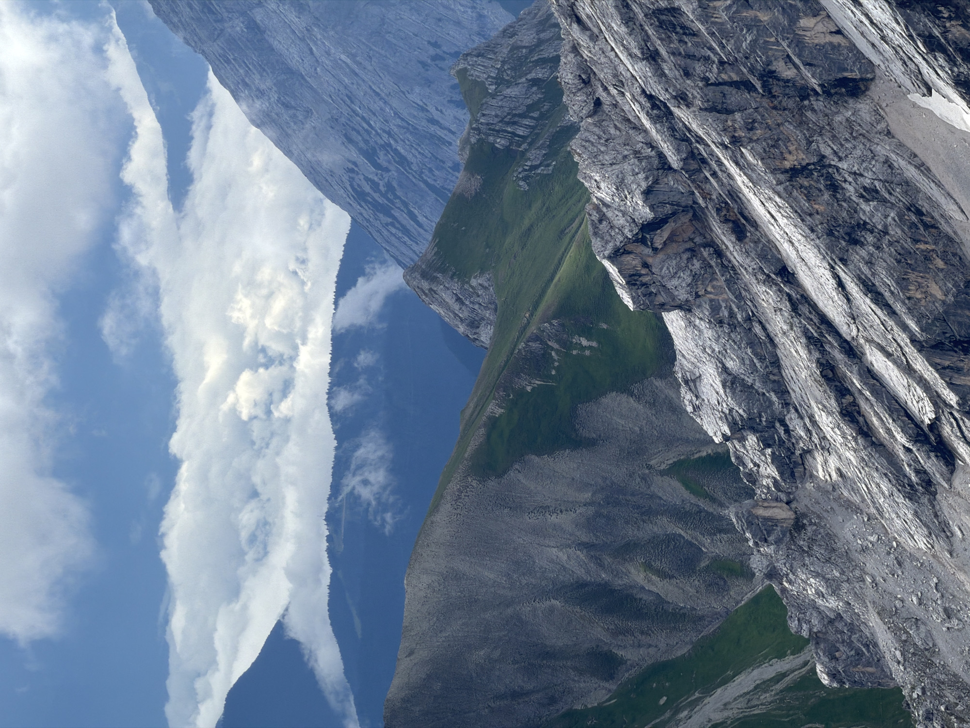 The image depicts a majestic mountain range under a partly cloudy sky. The foreground is dominated by a rugged, rocky slope with visible strata and debris. Behind this, a gentler, grass-covered mountain slope creates a striking contrast. Further in the distance, several layers of mountains recede, with the furthest peaks appearing hazy and bathed in a soft, blue light. Wisps of clouds cling to some of the mountain slopes, adding depth and atmosphere. The scene is one of natural grandeur, with a focus on the textures and geological formations of the mountains. This is likely a view of the Alps, possibly near Garmisch-Partenkirchen, Germany, given the characteristic appearance of the Bavarian Alps. There are no people or man-made objects immediately visible, emphasizing the raw, untamed nature of the environment.