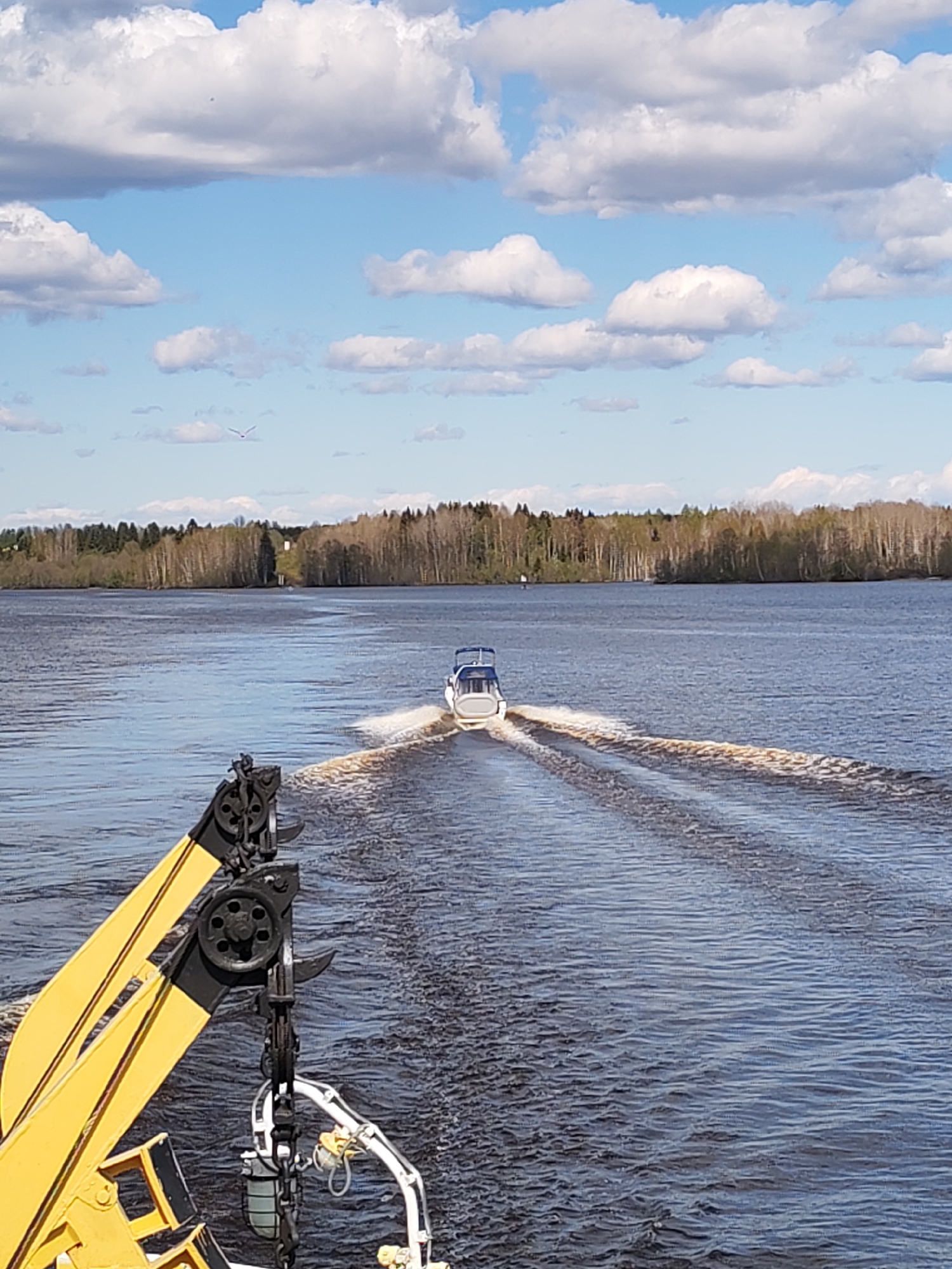 The image shows a body of water with a boat moving away from the camera, leaving a wake behind it. The boat is small and white with a blue canopy. The water is dark and choppy. In the foreground, there is a yellow and black crane-like structure. In the background, there is a line of trees on the opposite shore. The sky is blue with scattered white clouds. The picture was taken at Kondratovo, Russia.