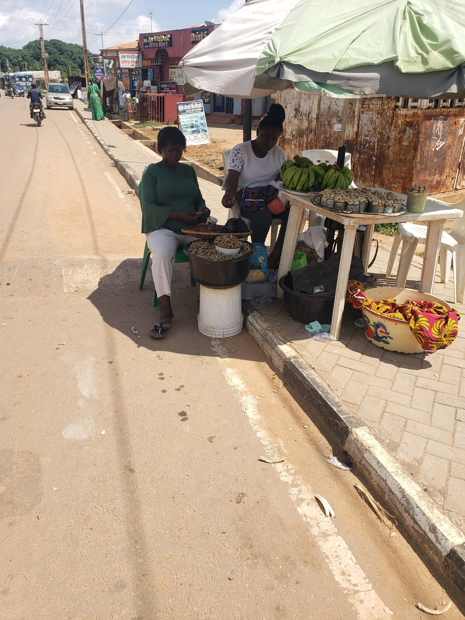 Two women are sitting at a roadside stall selling bananas and peanuts in what appears to be a sunny, outdoor urban environment. The stall has a white plastic table covered with a light green and white umbrella. On the table, there are bunches of green bananas and several trays filled with peanuts. One woman, wearing a dark green long-sleeved shirt and white pants, is seated on a green plastic chair and is holding a small item in her hands, possibly sorting peanuts. The other woman, wearing a white short-sleeved top and dark pants, is also seated and looking at the peanuts. In front of the stall, there are several large containers, including a black plastic tub and a white bucket, also containing peanuts. To the right of the table, a large, colorful patterned fabric is draped over a bowl. The ground is paved with interlocking bricks and a dirt shoulder. In the background, there are buildings, including a shop labeled "JervidDar Ultra Mart", and trees. A motorcycle and a car are visible on the road to the left of the scene. The overall impression is of a busy street-side market.