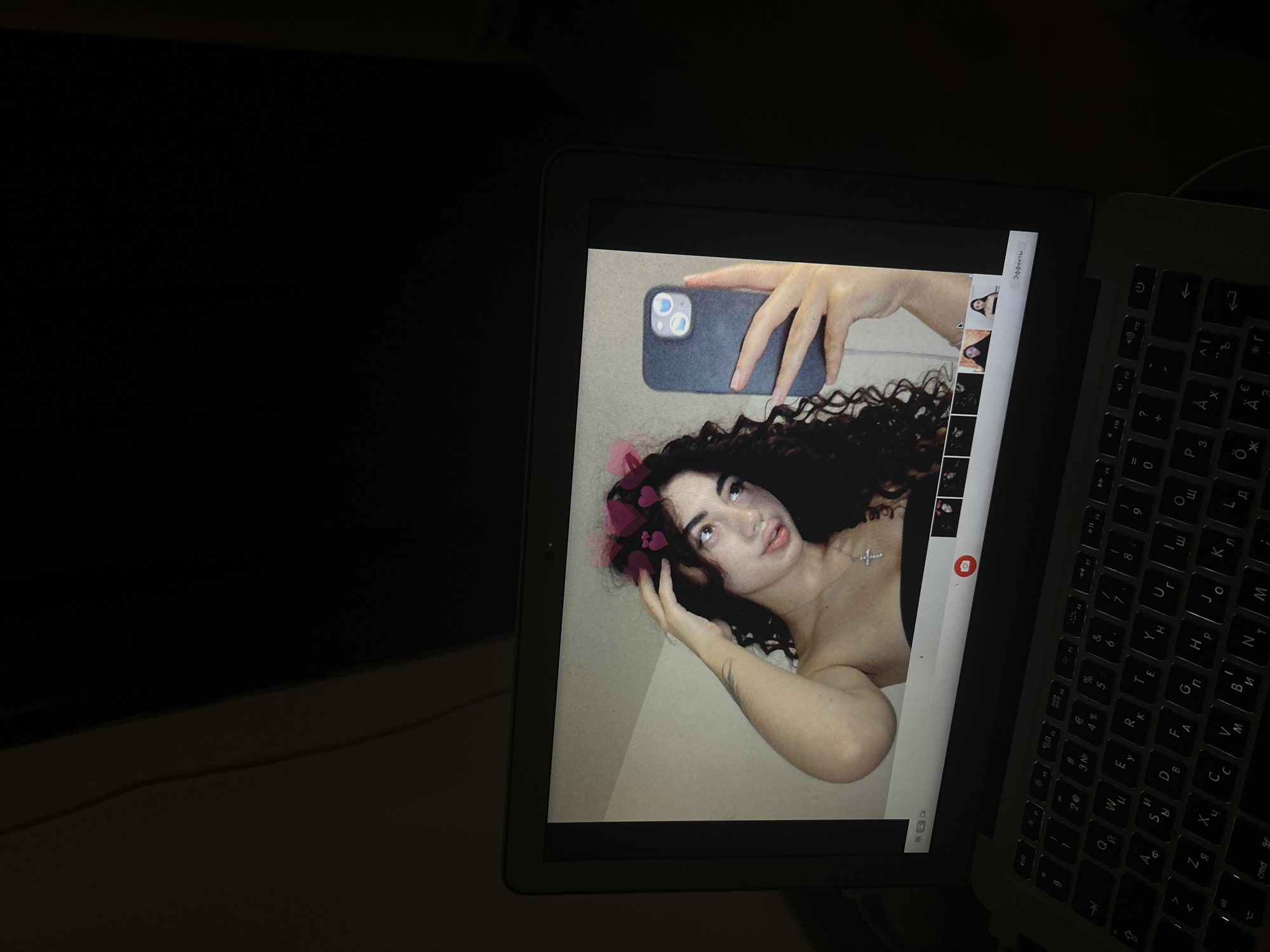 A laptop displays a selfie of a young woman with curly dark hair, wearing a black strapless top and a silver cross necklace. She has pink heart filters over her head and is holding a dark-colored smartphone in her right hand, taking the picture. The laptop is on a dark surface, and the surrounding environment is dimly lit, suggesting an indoor setting. The image does not contain any information about the city of Chotěšov, Czechia.