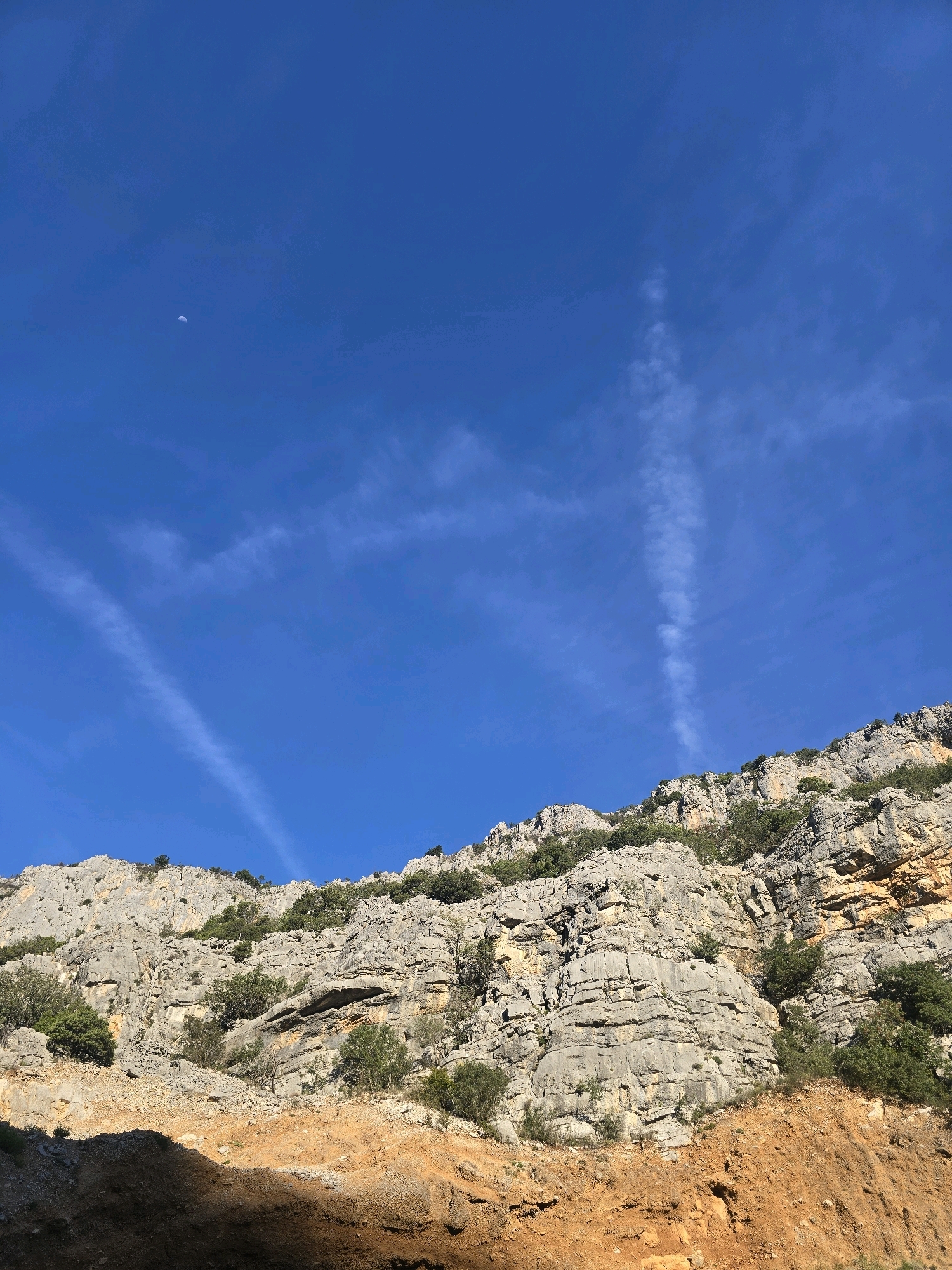 The image depicts a natural landscape with a prominent rocky mountain face under a clear blue sky. The mountain is layered with various shades of grey rock and interspersed with patches of green vegetation. The foreground features a steep, reddish-brown embankment. Several wispy contrails are visible in the sky, suggesting airplane activity. A small, crescent-shaped moon is also visible in the upper left portion of the sky. The scene appears to be a rugged, undeveloped area, consistent with a mountainous region. While the image is described as being in Podgorica, Montenegro, there are no urban elements or recognizable landmarks to confirm this specific location. The image is a still photograph with no discernible action or human subjects.