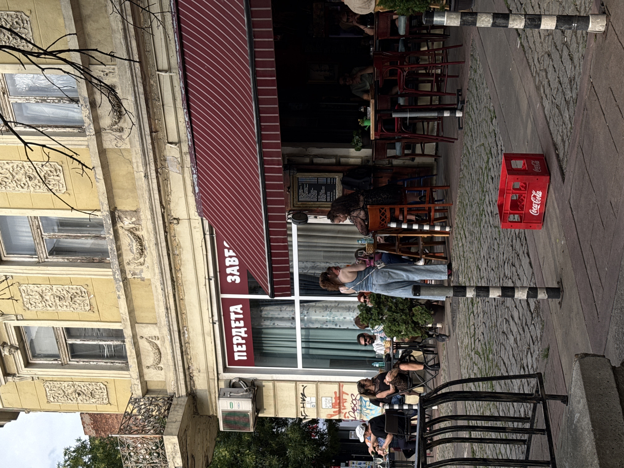 An outdoor seating area of a cafe or restaurant is depicted in the city of Sofia, Bulgaria. The facade of the building is yellow with ornate decorative elements around the windows and along the upper facade. A red and white striped awning covers the outdoor seating area, which features tables and chairs. People are seated at several tables, some conversing, others looking around. A woman in light blue overalls stands and faces away from the camera. In the foreground, on the cobblestone pavement, a red Coca-Cola crate is visible. The street is paved with cobblestones, and a black and white striped bollard stands to the right of the crate. Trees are visible on the left side of the image, and the sky is overcast.