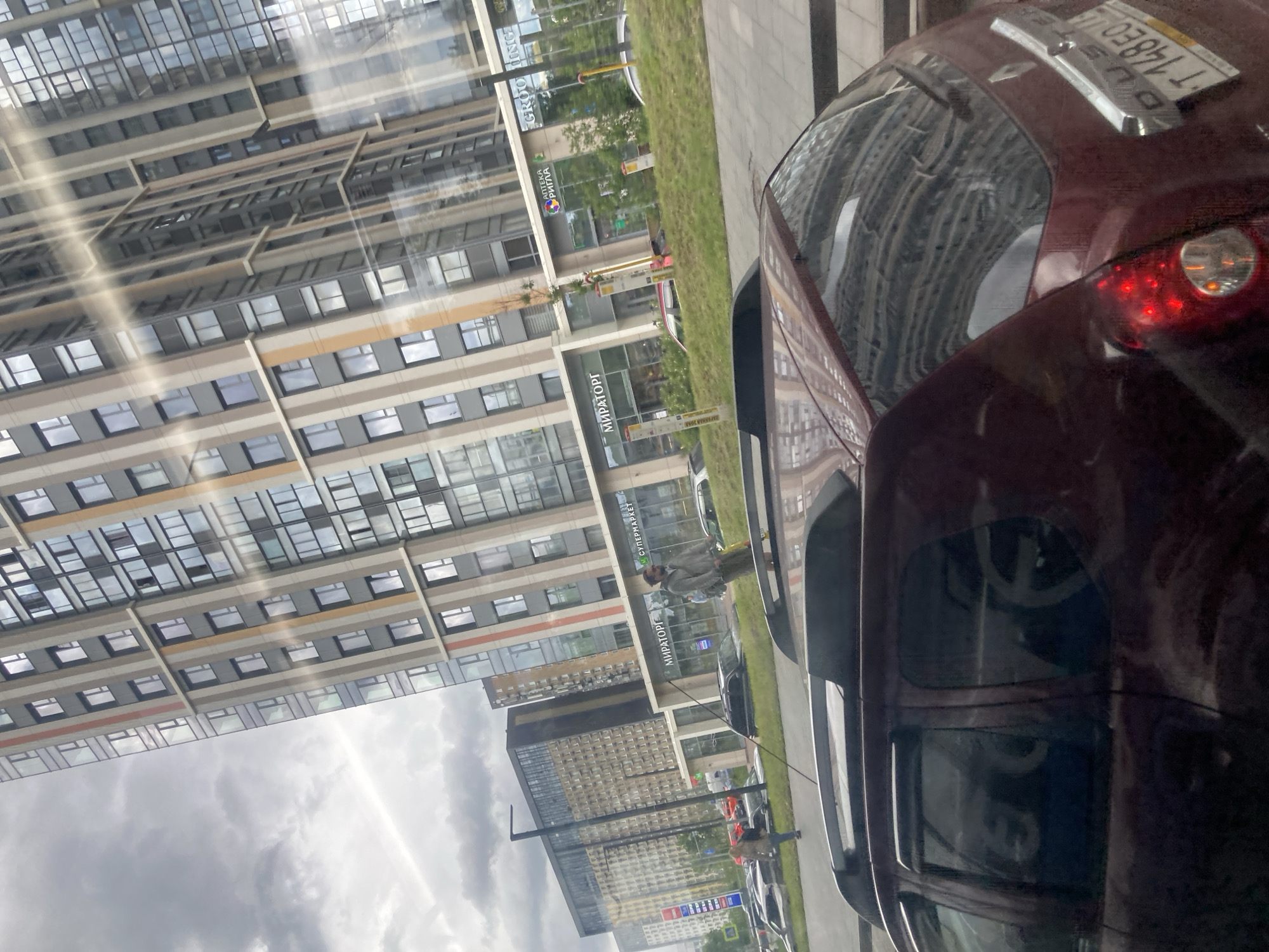 The image captures a scene in what appears to be a modern urban environment, possibly Moscow, Russia, as indicated by the license plate visible on the maroon car in the foreground. The license plate reads "T 148EO", where EO is likely the Cyrillic letters ЕО.
Dominating the view is a tall building with a modern architectural style, featuring numerous windows. The reflection in the car's window suggests that this building is across the street or directly adjacent to the viewer.
On the street level, visible through the car's windows, there's evidence of a supermarket or a similar commercial establishment identified by the inscription "МИРАТОРГ" (MIRATORG), a well-known Russian meat processing company and supermarket chain. Other storefront signs are also visible, with the word "АПТЕКА" (Pharmacy) present, and there are other businesses.
People are present on the sidewalk in front of the building. The scene implies a typical urban pedestrian and commercial setting. A patch of green suggests a landscaped area, separating the sidewalk from the road.