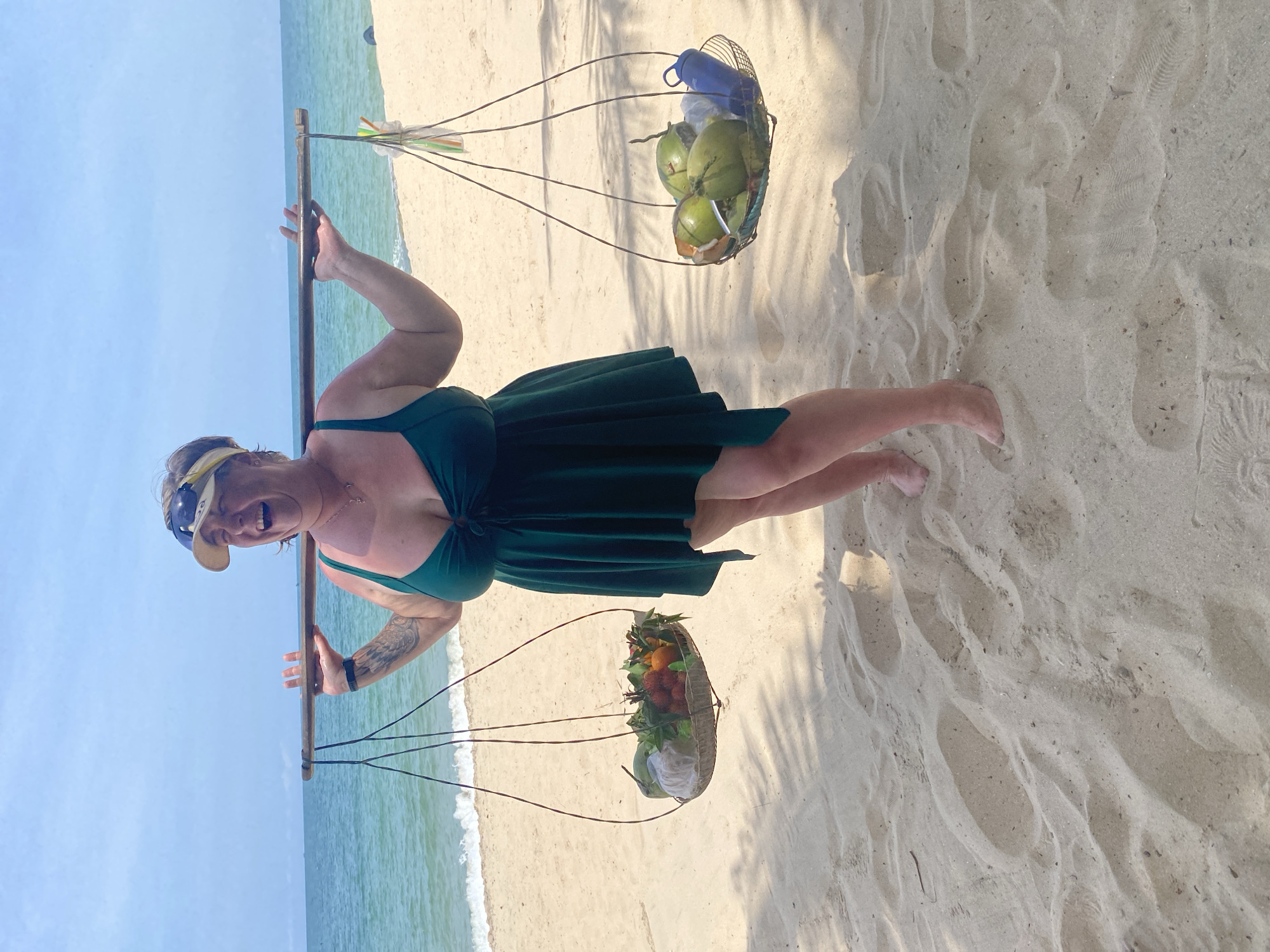 A woman is standing on a sandy beach in Phu Thuy Ward, Vietnam, carrying a yoke across her shoulders with two baskets of fruit and coconuts. She is wearing a green one-piece swimsuit and a yellow visor. The ocean is visible behind her, with gentle waves lapping at the shore. The sky is clear and blue. The sand is light brown and appears to be dry, with some footprints visible. The baskets contain various fruits, including rambutans, oranges, and what appear to be lychees, as well as several coconuts and a blue water bottle. The woman is smiling broadly, with her mouth open, and has her eyes closed. She has tattoos on her left arm and is wearing a black fitness tracker on her left wrist. The image captures a moment of enjoyment and relaxation on a tropical beach.