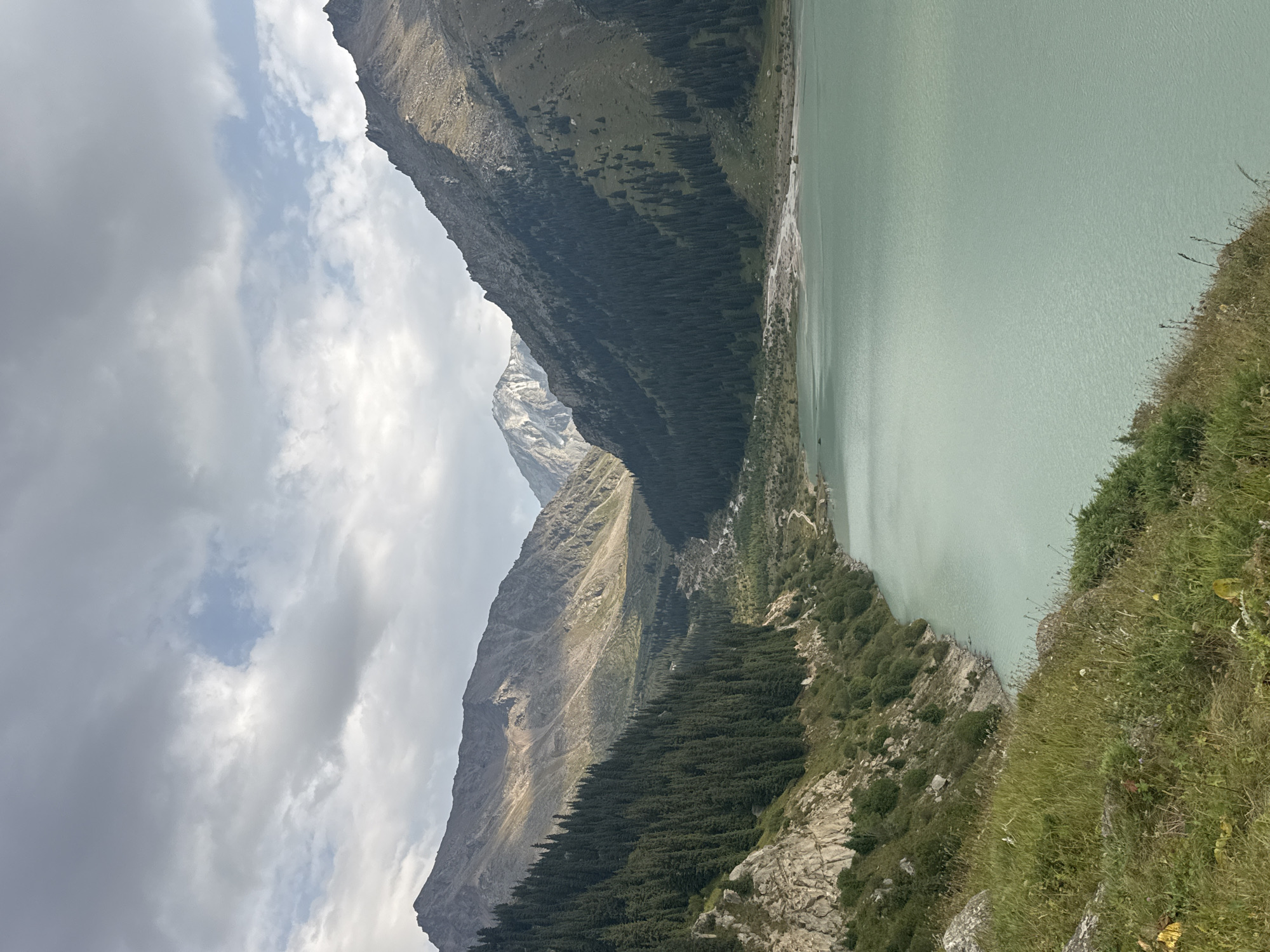 This image depicts a scenic mountainous landscape with a large, placid lake in the foreground. The lake's water is a light turquoise color. Steep, tree-covered slopes rise on either side of the lake, transitioning into rugged, rocky mountains in the background. The sky is partially overcast with dramatic clouds. A winding dirt road is visible along the shoreline in the mid-ground. The overall impression is one of natural grandeur and remote beauty. This location is likely Big Almaty Lake, a popular natural landmark located in the Trans-Ili Alatau mountains south of Almaty, Kazakhstan. There are no people or discernible actions in the image; it is a static landscape photograph.