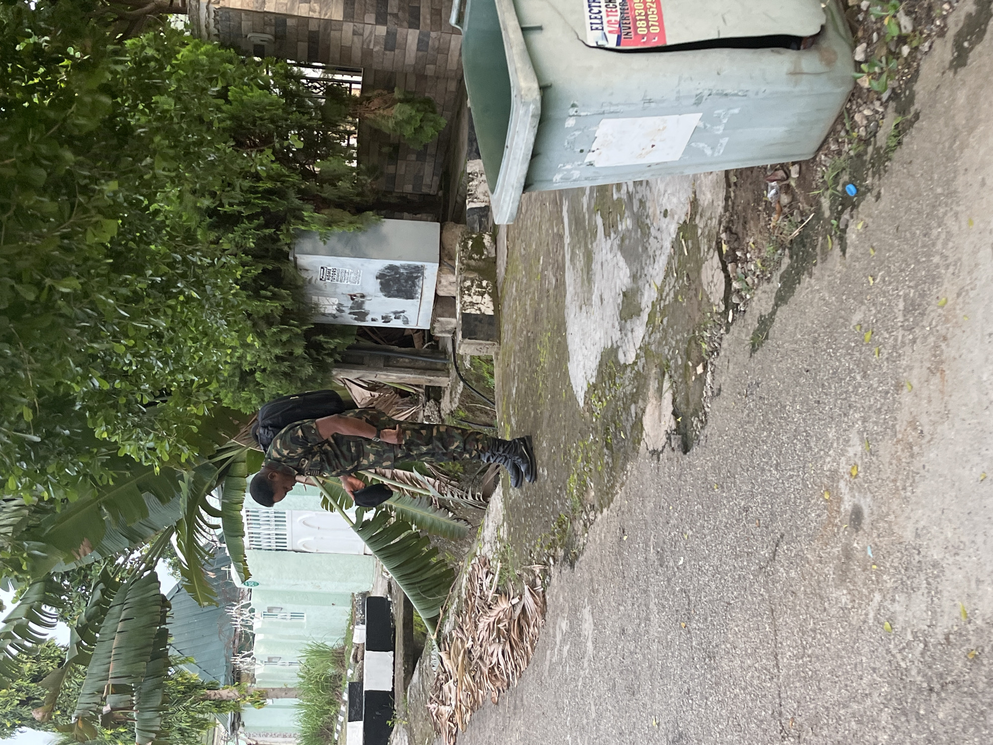 A Black male dressed in camouflage military fatigues, a black backpack, and black boots stands on a paved area next to a street. He is bending over, holding a black cap in his left hand, and appears to be interacting with a large, partially defoliated banana plant that extends from a ditch or channel. The street is asphalt and shows signs of wear and some standing water. To the right of the scene is a large, pale green plastic trash receptacle with some labeling that is partially obscured. Behind the trash receptacle, a brick wall is visible. In the background, a light green wall with white trim and white gates can be seen, with lush green foliage and trees surrounding it. A metal utility box with some posters attached is located in the mid-ground behind the banana plant. The overall environment suggests an outdoor urban or suburban setting in Abuja, Nigeria, possibly near a residential or commercial area with significant vegetation.