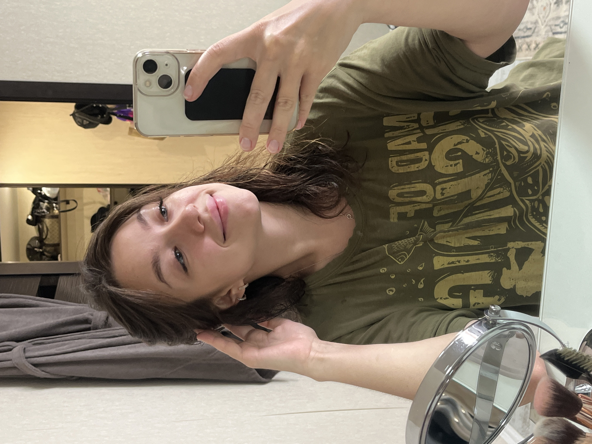 A young woman with medium-length brown hair is taking a mirror selfie. She is wearing a dark green t-shirt with a yellow graphic design on it. Her left hand is raised, touching her hair near her temple, and her right hand is holding a smartphone. She is smiling gently, looking at her reflection. In the background, a gray bathrobe hangs on a hook, and shelves with various items are visible. A makeup mirror and brushes are in the foreground. The location is identified as Chaikovsky, Russia.