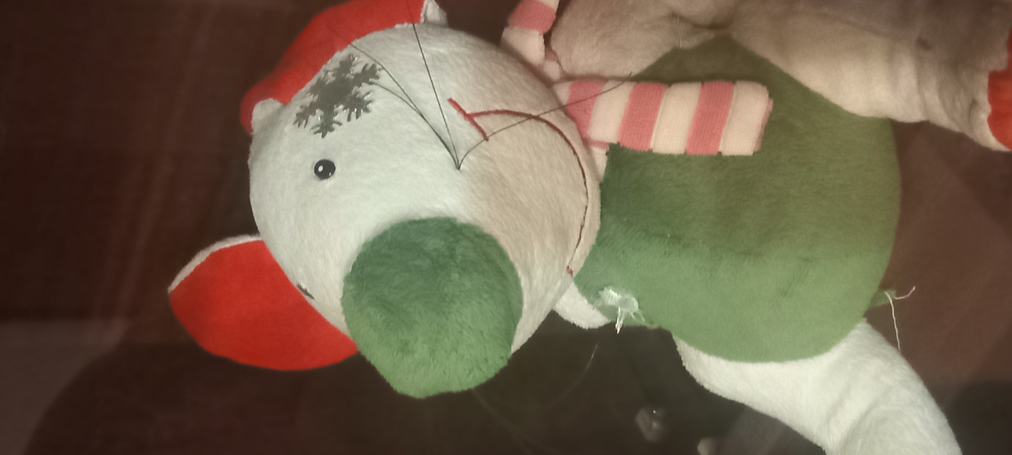 A stuffed toy mouse is shown in a close-up, vertically oriented, medium shot. The mouse is predominantly white with a large green nose, red ears, and a green body. It wears a red and white striped scarf. A small snowflake-like embellishment is affixed to its forehead, and black thread whiskers extend from its snout. A single black eye is visible. The background is dark and indistinct. No location in Kryvyi Rih, Ukraine, can be determined from the image.