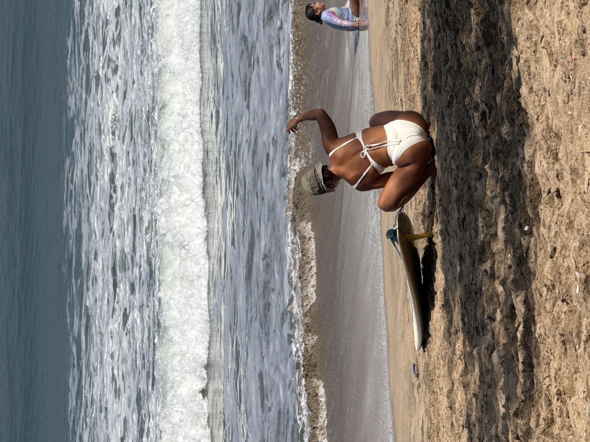 The media file shows a beach scene at Kuta, Indonesia. The image features two people and a surfboard. One person, a woman in a white bikini, is crouched on the sand next to a surfboard, appearing to be in motion or preparing to move. She is wearing a hat and her back is to the camera. Another person, a child wearing a multicolored long-sleeved swimsuit, is lying on the sand closer to the water.
The scene depicts the edge of the ocean with waves breaking on the shore. The sand is visible, showing textures and footprints, suggesting recent activity. The light suggests it is daytime, possibly late afternoon due to the angle of the shadows. The weather appears to be clear and sunny. The background shows the ocean and the sandy beach, typical of a tropical location like Kuta. There is no visible text in the media file.