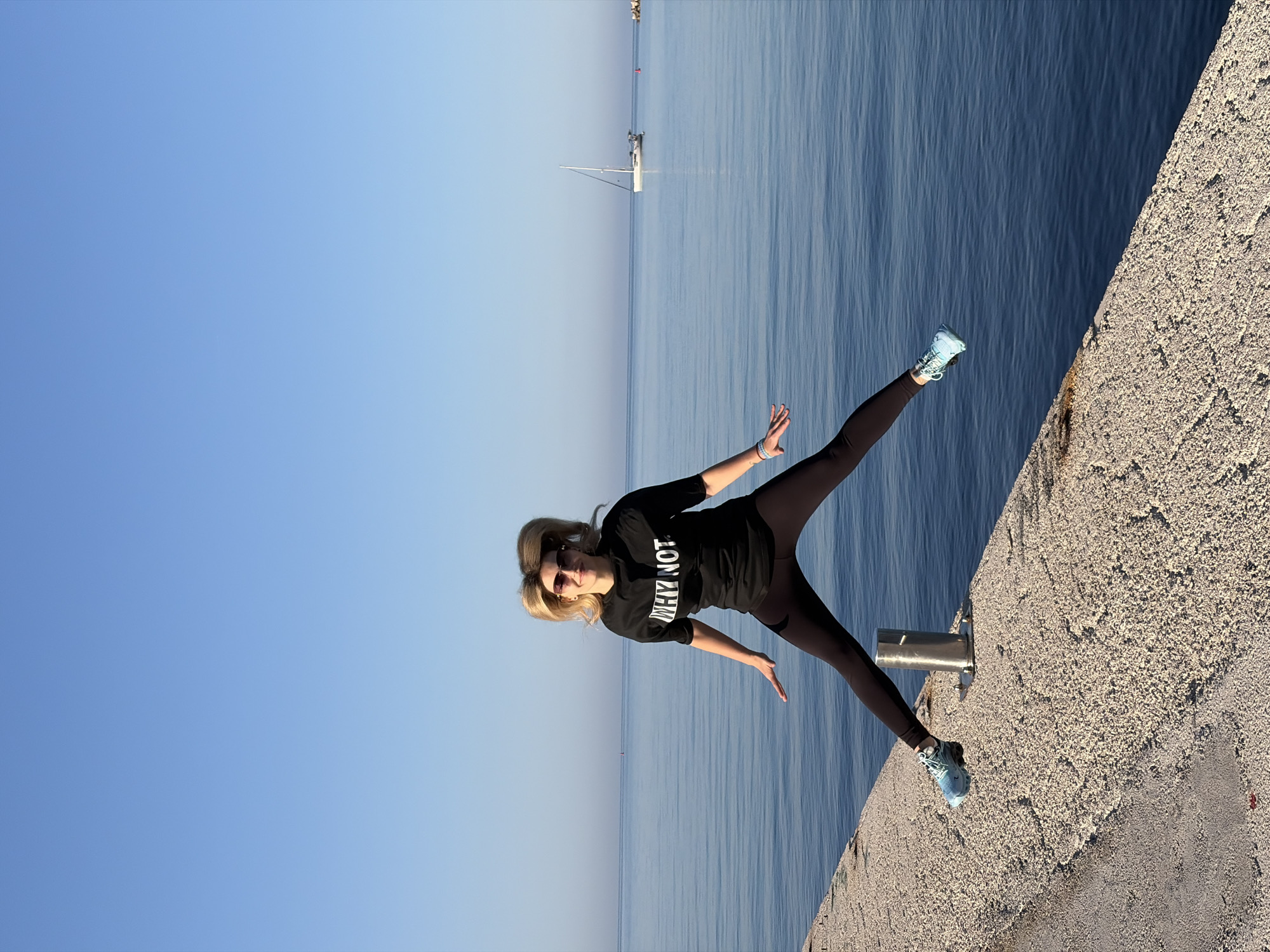 A full-body shot of a female subject is captured on what appears to be a concrete breakwater. The woman is wearing dark athletic leggings, a black, long-sleeved shirt with "WHY NOT" printed in bold, white letters, and light blue athletic shoes. She has short blonde hair and sunglasses on her head. Her posture suggests she is in motion, with arms outstretched and legs in a split position, one on the edge of the breakwater and the other extended out over the water.
The background features a flat, calm expanse of sea and a cloudless, light blue sky. A single sailboat is visible in the distance on the water. The concrete breakwater is textured with small, rough aggregate, and has a small, cylindrical metal object affixed to it.
Based on the visual elements of the landscape, the location appears to be in Budva, Montenegro, along the Adriatic coast.
