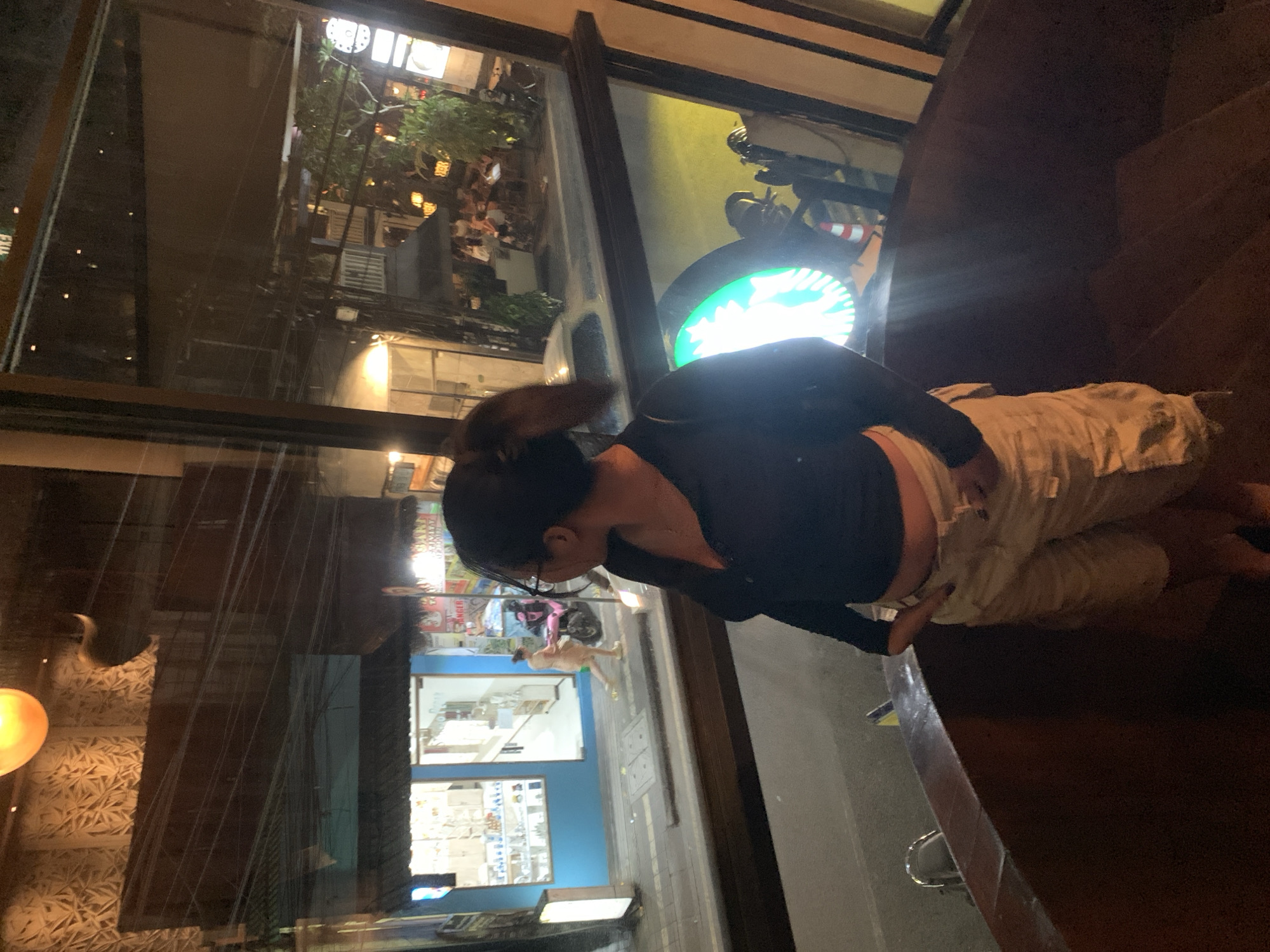 The image depicts a woman sitting at a wooden bar inside a building, possibly a cafe or restaurant, at night. She is wearing a dark long-sleeve shirt and light-colored pants. The background shows a street scene through a large window. In the window's reflection, one can see signs, lights, shops, and people walking on the sidewalk. There is a Starbucks logo visible outside. The location is likely the Ubud District, Indonesia.