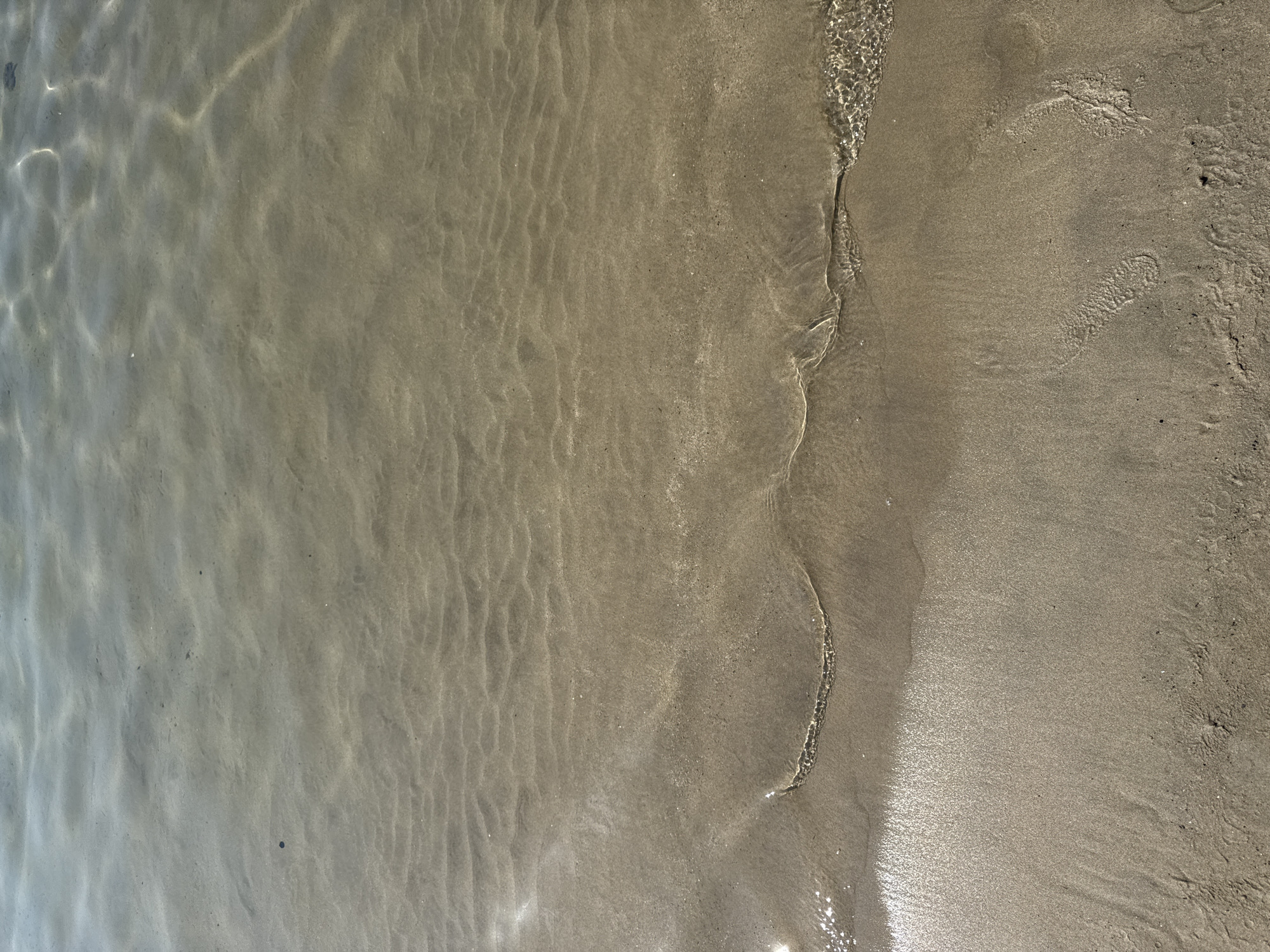 The image shows a close-up, high-angle view of the shallow water at the edge of a sandy beach. The water is clear and calm, with sunlight glinting off the surface, creating ripples and light patterns on the sand beneath. The sand is fine-grained and a light brown color, showing subtle undulations from the water's movement. A thin line of receding water marks the current shoreline. The scene depicts a tranquil coastal environment, characteristic of beaches in Sanremo, Italy. There are no people or distinct objects visible in the frame, only the natural elements of water and sand.