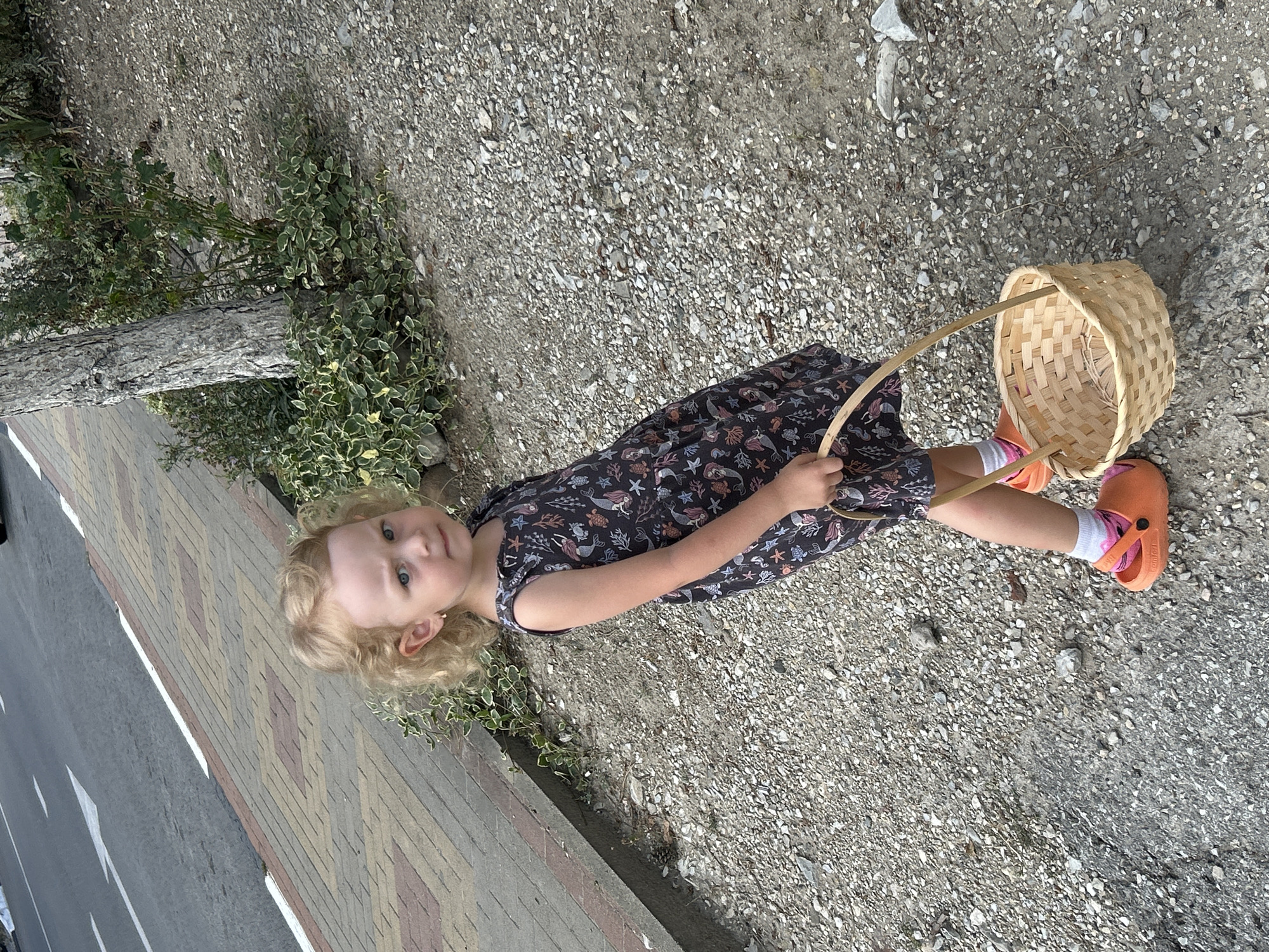 A young girl with curly blonde hair, wearing an orange crocs, white socks, and a dark dress with a marine print, stands on a gravel path. She holds a small wicker basket in her right hand. The background shows a paved sidewalk, a road with a white arrow marking, and some vegetation. The scene appears to be on a public street or walkway, possibly in a coastal area like Gelendzhik, Russia, given the presence of a road, sidewalk, and sparse vegetation.