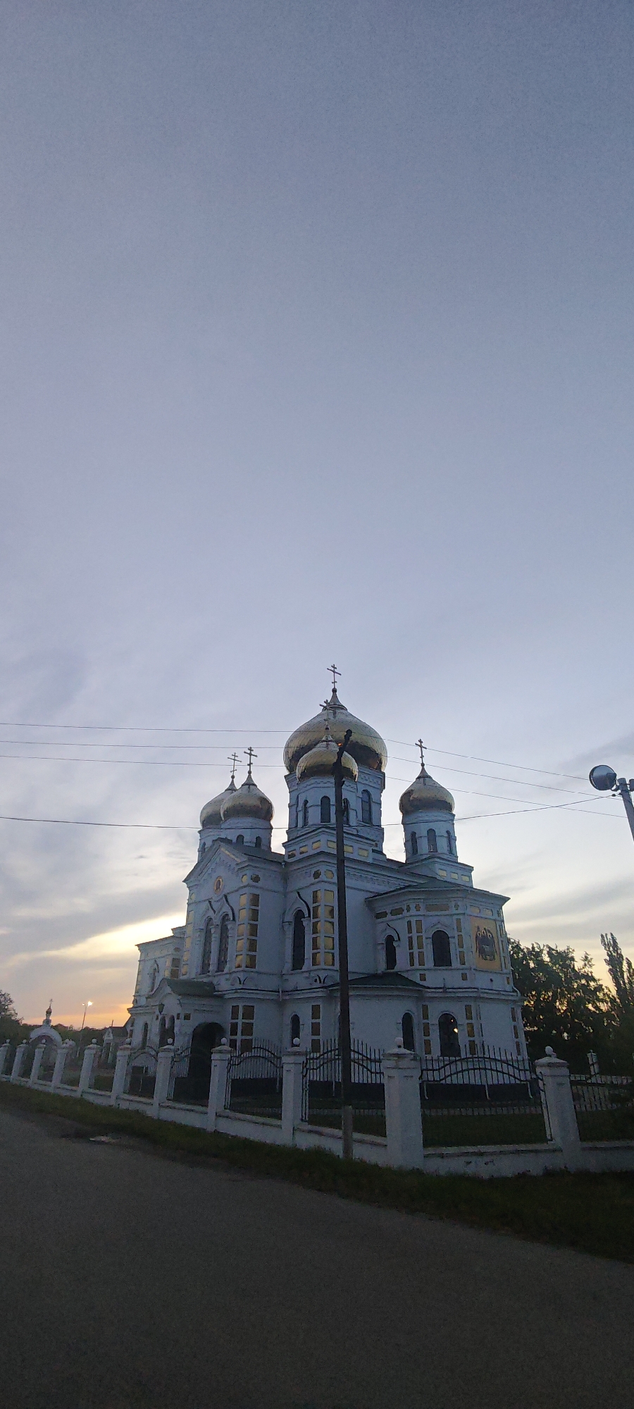 A low-angle, eye-level shot depicts the Cathedral of St. Nicholas the Wonderworker in Novodonetskaya, Russia. The cathedral, constructed of light-colored material, features prominent golden domes topped with crosses. The sky is pale blue, tinged with hues of dusk or dawn. Power lines stretch horizontally across the frame, intersecting the top portion of the building. A streetlight is seen to the right. A low, white fence with black metal railings surrounds the cathedral grounds. The street and a small strip of grass verge are visible at the bottom of the frame.