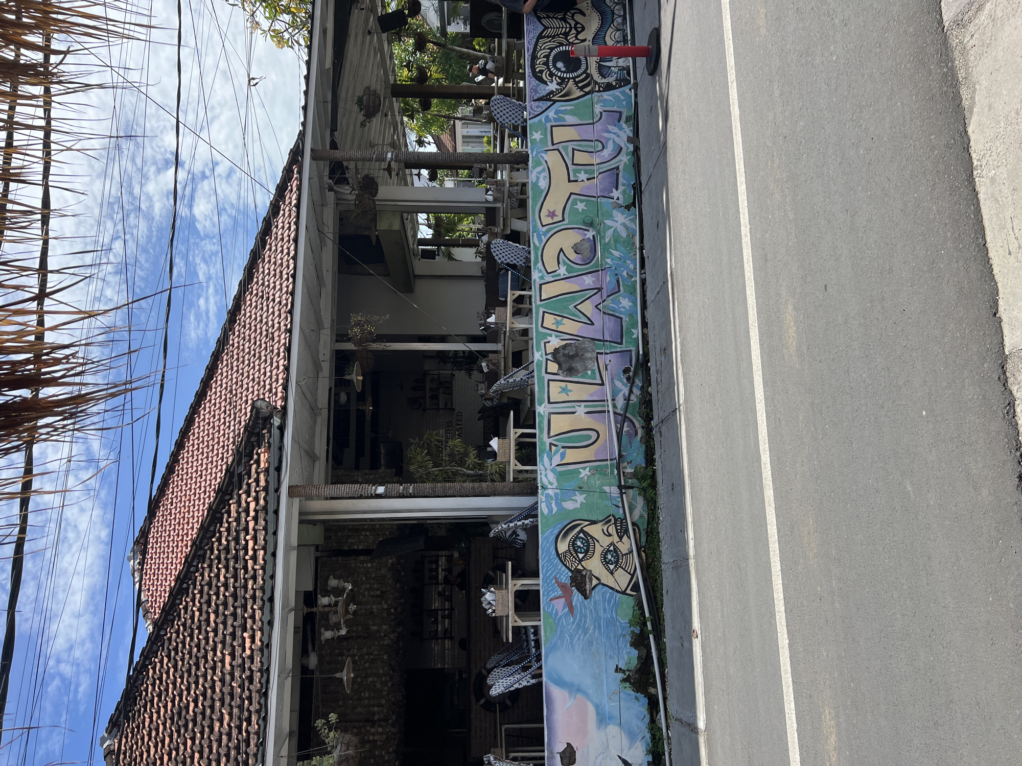 The image depicts a two-story building in Kuta Selatan, Indonesia, with a red-tiled roof partially visible against a blue sky with scattered clouds. The building appears to be a restaurant or cafe, featuring open-air seating areas with tables and chairs on both levels. A colorful graffiti-covered wall or barrier runs along the side of the building, adjacent to a paved road with a white lane marker. The graffiti includes lettering and depictions of faces or figures. The overall environment suggests a daytime urban scene.