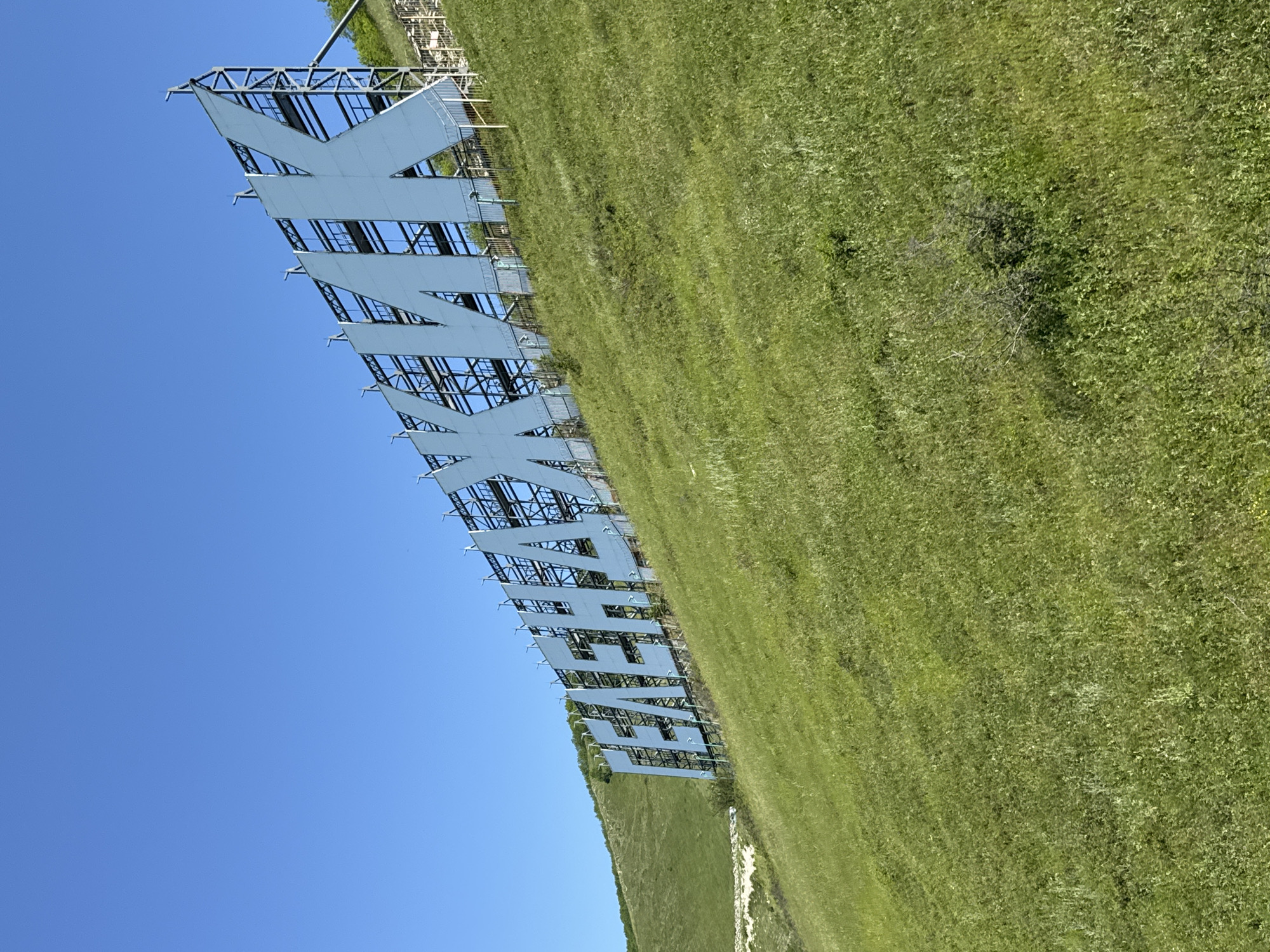 The image displays a large, prominent sign spelling out "ГЕЛЕНДЖИК" in capital Cyrillic letters, situated on a grassy hillside under a clear blue sky. The letters are white and three-dimensional, supported by an exposed metal framework that provides structure and depth. The sign extends horizontally across the frame, positioned above a slope covered in dense, green grass.
The scene depicts an outdoor environment, bathed in bright daylight, indicative of a clear, sunny day with no clouds visible in the expansive blue sky. No human activity or people are present, placing the focus entirely on the monumental sign and the natural landscape. The grass appears well-maintained, suggesting a public or landmark area. The context indicates the location is Gelendzhik, Russia, which is directly spelled out by the sign itself. The overall impression is that of a landmark or welcome sign in a picturesque, natural setting.
