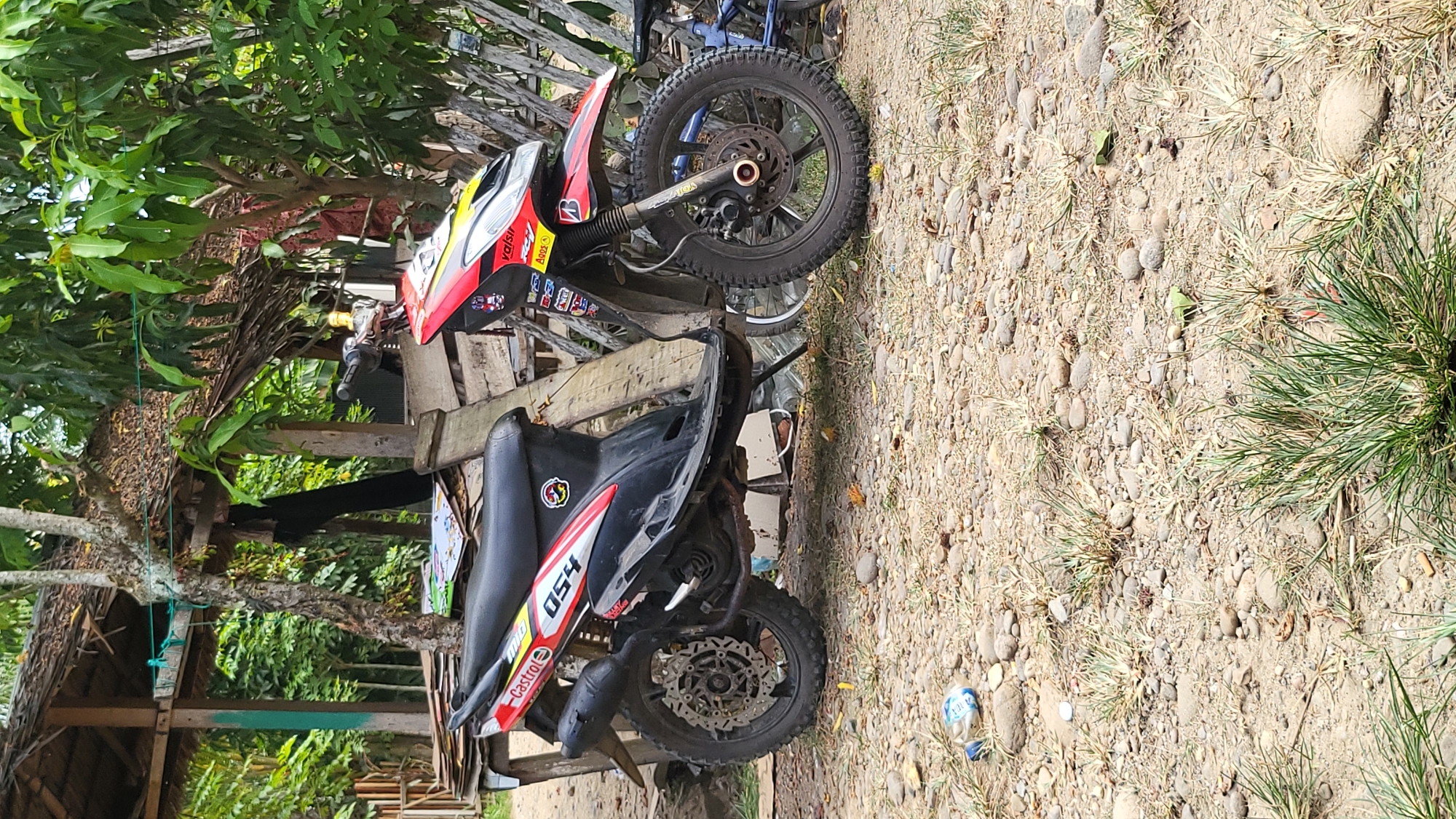 The image shows two motorcycles parked outdoors on a dirt and gravel surface. The motorcycles are the primary subjects, with one being a red and yellow dirt bike and the other a black and red scooter. The dirt bike is positioned in the background, leaning against some wooden structures. It features knobby tires, typical for off-road riding. The scooter is in the foreground, also parked on the ground.
The setting appears to be a rural or semi-rural area, indicated by the natural ground cover, sparse vegetation, and what looks like a simple wooden shelter or structure behind the motorcycles. Lush green foliage from trees is visible, suggesting a warm climate. The lighting suggests it is daytime, likely a sunny day given the clear shadows and bright illumination.
There is some visible text on the motorcycles. The red and yellow dirt bike has racing decals, including "val-sil" and what appears to be a logo. The black and red scooter has the number "054" prominently displayed, along with the "Castrol" brand name in red text. A partially visible plastic bottle is on the ground near the scooter. No people are present in the image. The overall impression is one of stillness, with the motorcycles parked and unattended in an outdoor environment. The location context is Teumpokteungku, Indonesia.