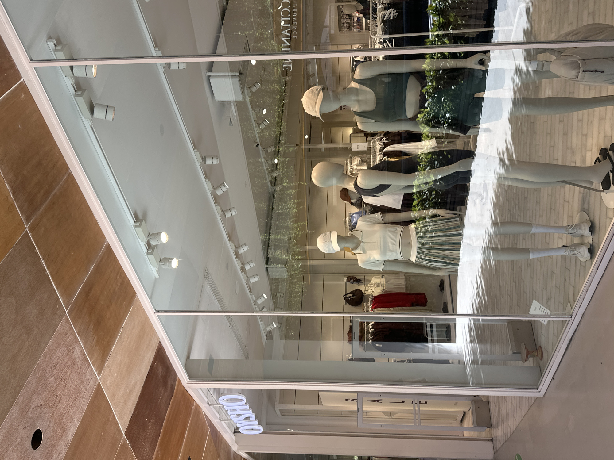 In the city of Kuta, Indonesia, a retail store display window showcases three mannequins in athletic wear. The store's logo, "OYSHO," is visible above the entrance. The mannequins are positioned in front of racks of clothing and decorative plants. The left mannequin wears a white collared shirt and a striped skirt. The middle mannequin is dressed in a dark tank top and shorts. The right mannequin is clad in a dark green sports bra and matching shorts. Reflections on the glass indicate an outdoor environment, possibly a shopping area. The interior lighting is a series of track lights, illuminating the merchandise.