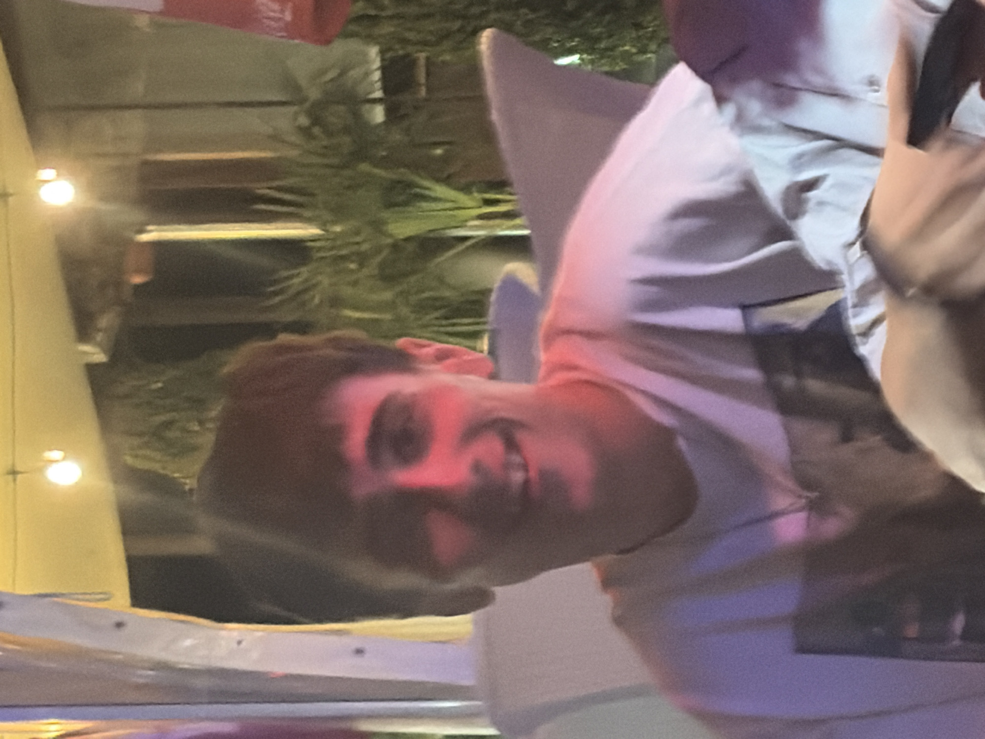 The image depicts an indoor or semi-outdoor nighttime scene, attributed to the city of Elhovo, Bulgaria. A young Caucasian male, approximately in his late teens or early twenties, occupies the central foreground, positioned from the chest up. He possesses short, dark hair and is smiling broadly, revealing his teeth, while looking directly at the viewer. A prominent red light source illuminates the left side of his face (viewer's right), his neck, and extends slightly onto his white, short-sleeved t-shirt. The t-shirt features a faded, dark graphic print on the chest, the details of which are indistinct. A silver-colored chain with a small, simple pendant rests around his neck, visible against the t-shirt.
In the background, multiple warm-toned, spherical incandescent light bulbs are visible hanging from an unseen overhead structure, casting yellow light and creating lens flares. Green foliage, consistent with potted plants or garden landscaping, is visible behind the subject. Vertical structural elements, possibly pillars or wall sections of a building, are also present. To the far left, a light-colored, possibly white or off-white, vertical structure with visible fasteners is partially visible. Partial views of other individuals or objects suggest a social setting; a light-colored sleeve of another person's clothing is visible in the lower right, and a red object with indistinct white markings (potentially text or a logo) is partially visible in the upper right corner. The overall ambiance is one of a casual, illuminated evening gathering.