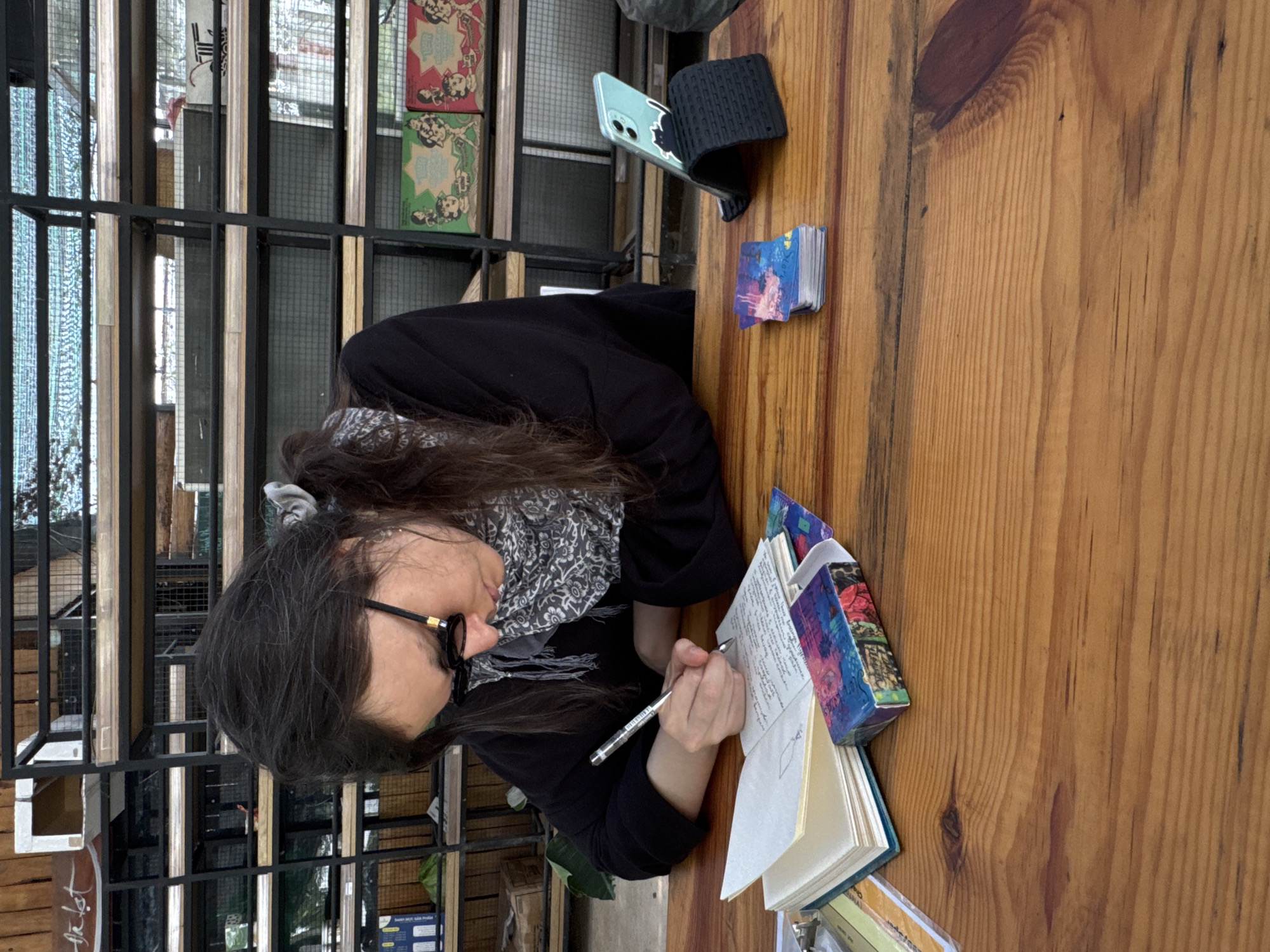 A woman with long, dark hair and glasses, wearing a black jacket and patterned scarf, is sitting at a wooden table. She is writing in an open notebook with a silver pen. A stack of small cards with colorful designs, a blue smartphone in a black case, and other books surround her. She is positioned near a window with a black metal frame, showing views of the outdoors and other buildings. The location is Da Lat, Vietnam, recognizable by the distinct architectural elements visible through the window.
