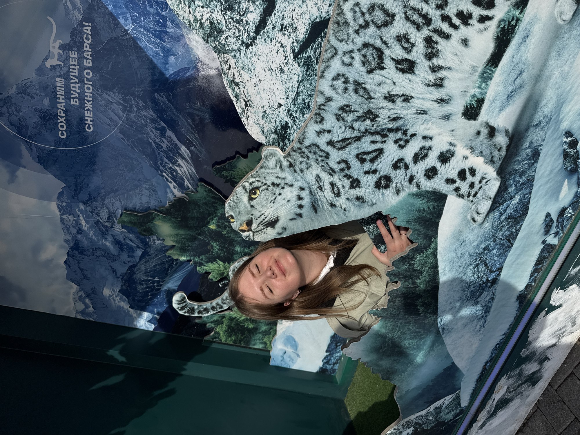 A photograph captures a young woman interacting with a public art display in Perm, Russia. The display is a depiction of a snow leopard and a mountainous backdrop. The woman is smiling with her eyes closed, seeming to pose or engage with the artwork. The artwork itself is brightly colored, combining the image of the snow leopard with a landscape scene. The artwork includes lettering in Russian "СОХРАНИМ БУДУЩЕЕ СНЕЖНОГО БАРСА!" (Save the future of the snow leopard!). The entire scene is set in an outdoor urban environment.