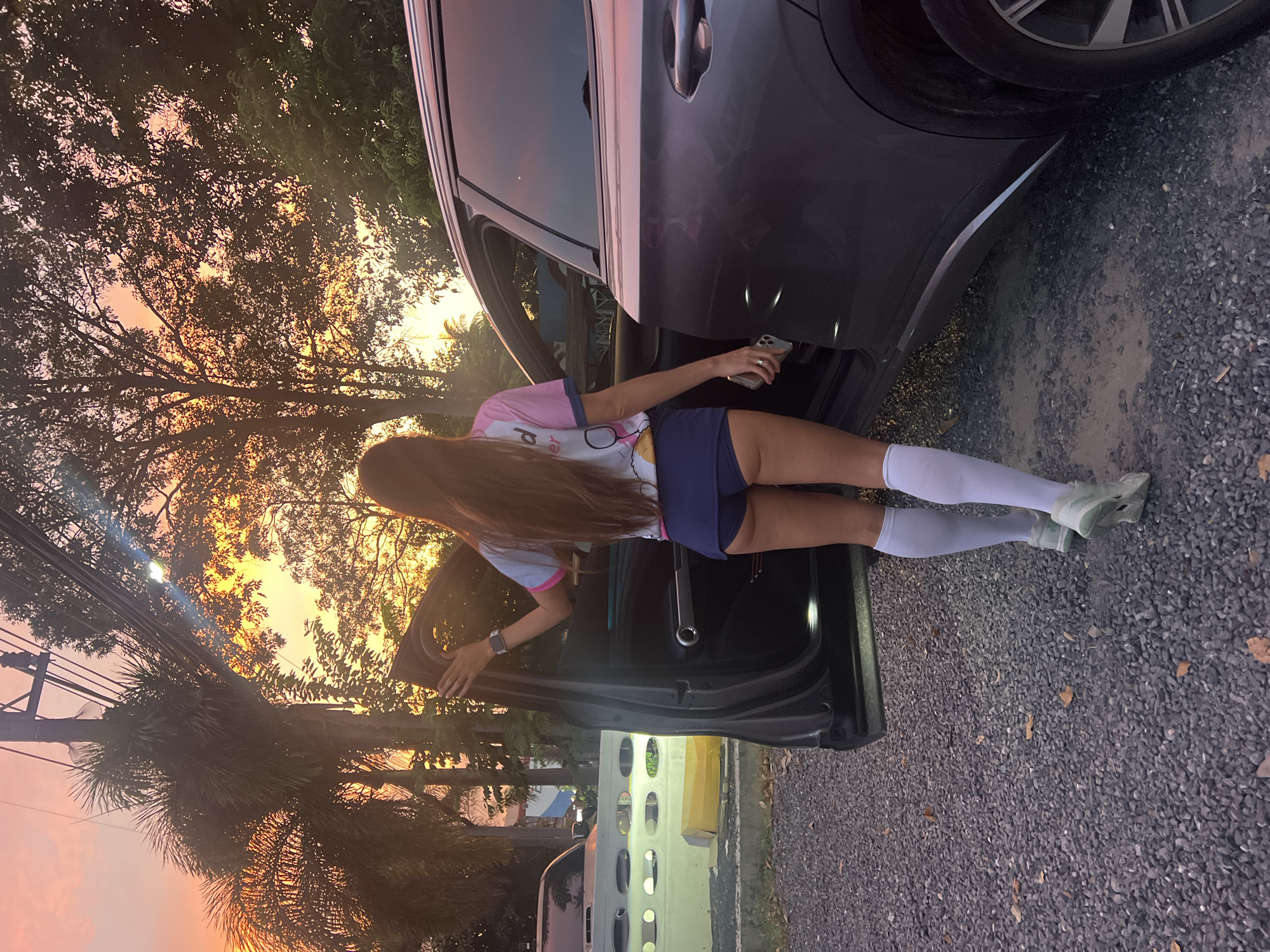 A full-body, rear-facing shot captures a woman with long, dark hair standing next to the open driver's side door of a dark gray SUV. The woman wears a pink and white t-shirt, navy blue shorts, white knee-high socks, and light green sneakers. She holds her phone in her right hand and her left hand rests on the open car door. The SUV is parked on a gravel driveway. In the background, the sky displays a colorful sunset with hues of orange, pink, and yellow, illuminating palm trees and other foliage. To the left of the SUV, a white car with illuminated circular lights is visible. The scene is set outdoors, with a wooden pole and overhead wires also present. The overall atmosphere is that of an evening scene in a tropical location.