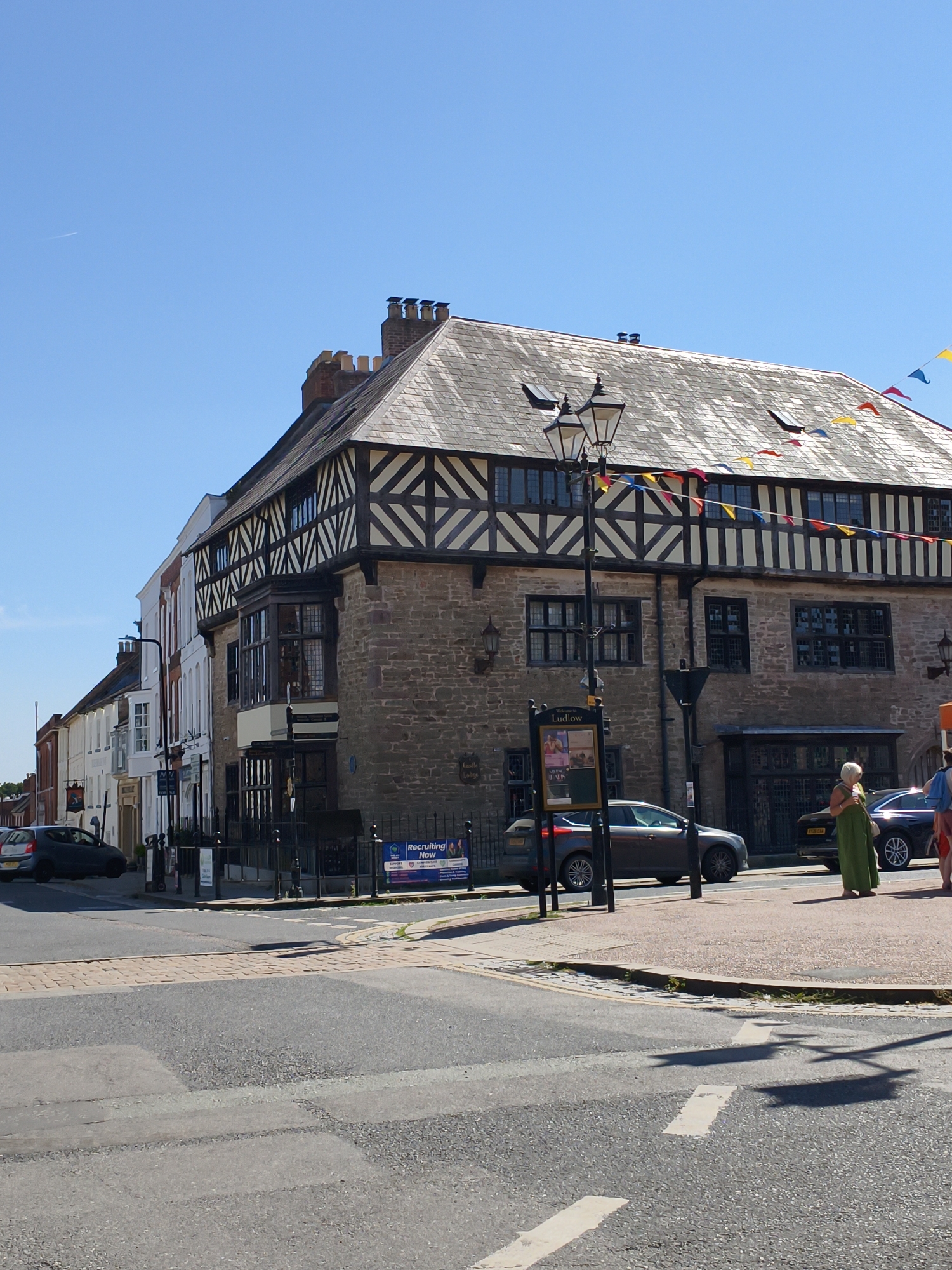 The image shows a bright, sunny day in Ludlow, United Kingdom. A historic stone and timber-framed building, likely an old inn or guildhall, dominates the right side of the frame. The building features characteristic Tudor-style dark timber beams against a pale stone facade, with multiple leaded glass windows. A steeply pitched tiled roof is visible, adorned with colorful triangular bunting strung across the upper levels. A black lamppost stands in front of the building, near a public information board. Several cars are parked on the street and on the road in front of the building, including a dark grey Ford Focus and a black saloon. A woman wearing a long green dress stands on the paved area in front of the building, looking towards the street. Another person in a pink top and blue shorts is partially visible to the far right. The street in the foreground shows a pedestrian crossing and road markings, indicating an intersection. In the distance, other buildings line the street, with a clear blue sky above. An airplane contrail is visible high in the sky.