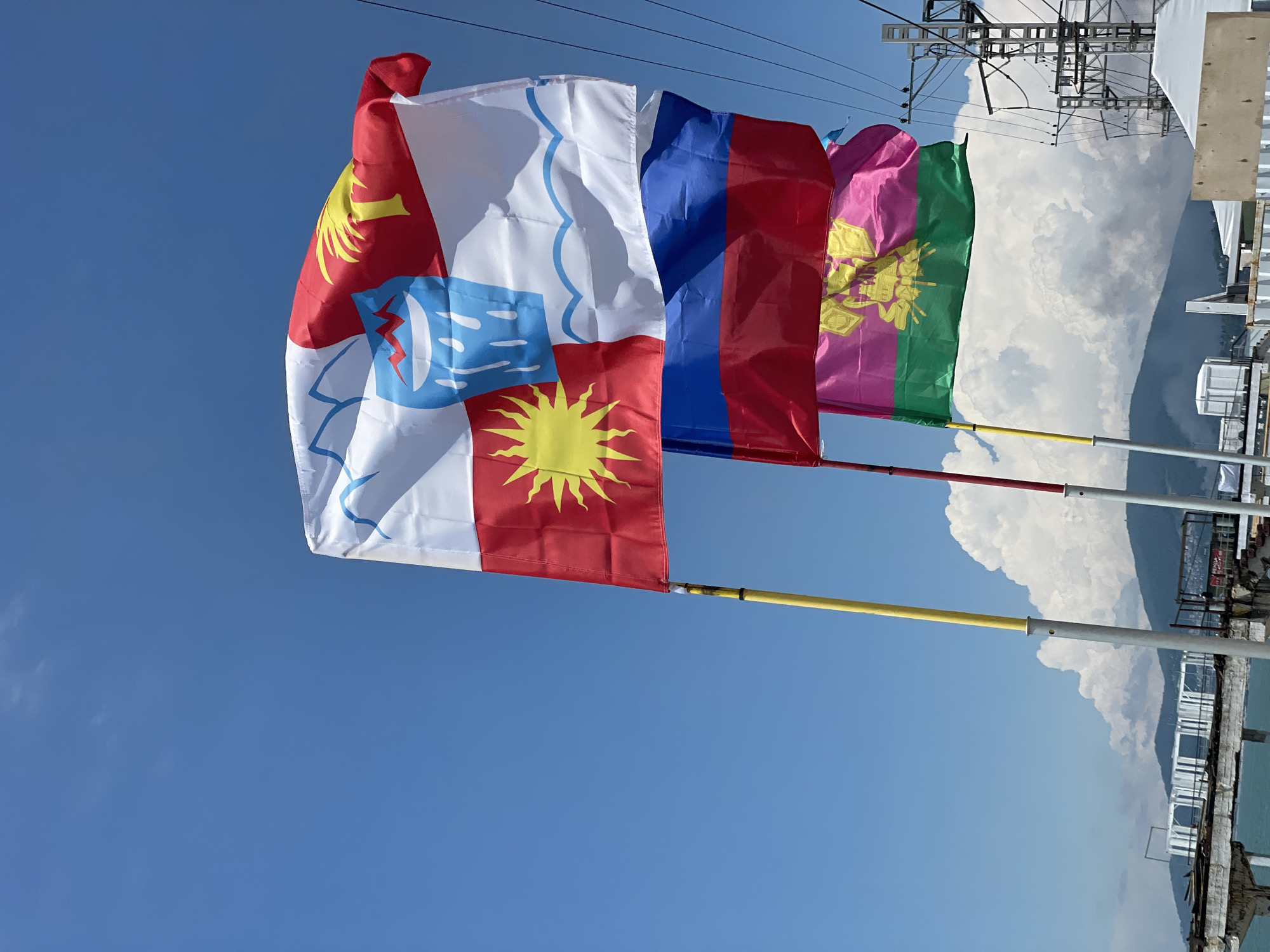 Three flags are displayed against a bright blue sky with scattered white clouds. The top flag is predominantly white with blue wavy lines at the bottom and a red section at the top right. This red section contains a yellow palm tree. A blue emblem with white lines representing water or rain is on the white part of the flag, next to a red section with a yellow sun. The middle flag is the flag of Russia, with white, blue, and red horizontal stripes. The bottom flag has a pink top half with a yellow emblem and a green bottom half. The flags are attached to poles, with yellow and red poles visible. In the background, there is a partially constructed pier or structure extending into the sea, with some industrial-looking buildings and power lines. The sea is visible on the left, and mountains can be seen in the distance. The overall setting appears to be a coastal area in Sochi, Russia.