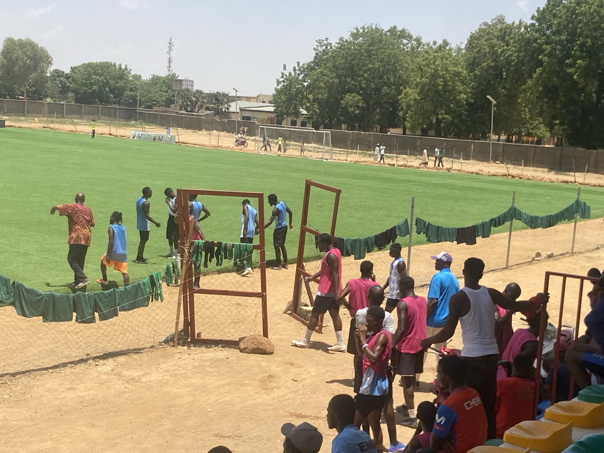 The image shows a soccer field in Tudun Wada, Nigeria, populated with young men. A group of young men, some in blue and black athletic wear and others in pink athletic wear, are on and near the edge of the soccer field. A man in an orange and brown patterned shirt is also walking near the field. Some green shirts are hanging on a chain-link fence. In the background, the soccer field is green. Other individuals can be seen walking on the field near the soccer goals. A fence surrounds the field and there are buildings and trees in the background. In the foreground, there are spectators seated in yellow and green stadium seating.