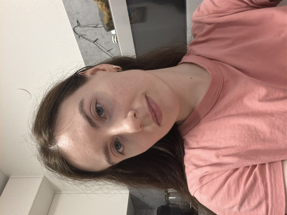 An individual is depicted indoors within what appears to be a kitchen setting in the city of Ковалёво, Russia. The subject is a female, seen from the mid-chest upwards, looking directly at the camera with a neutral expression. She has long, dark brown hair, fair skin, light-colored eyes, and is wearing a plain, coral-pink short-sleeved t-shirt. A small, gold-colored hoop earring is visible in her right ear. In the background, a white wall or ceiling is visible above grey tiled backsplash. A string of small, white LED fairy lights is affixed to the tiled wall. Below the lights, a light-colored appliance, possibly a microwave, is present, with a translucent, fluted bowl containing indeterminate contents resting on top of it. White overhead cabinets are partially visible in the upper left. Stacked dark-colored objects are partially visible in the far-left background.