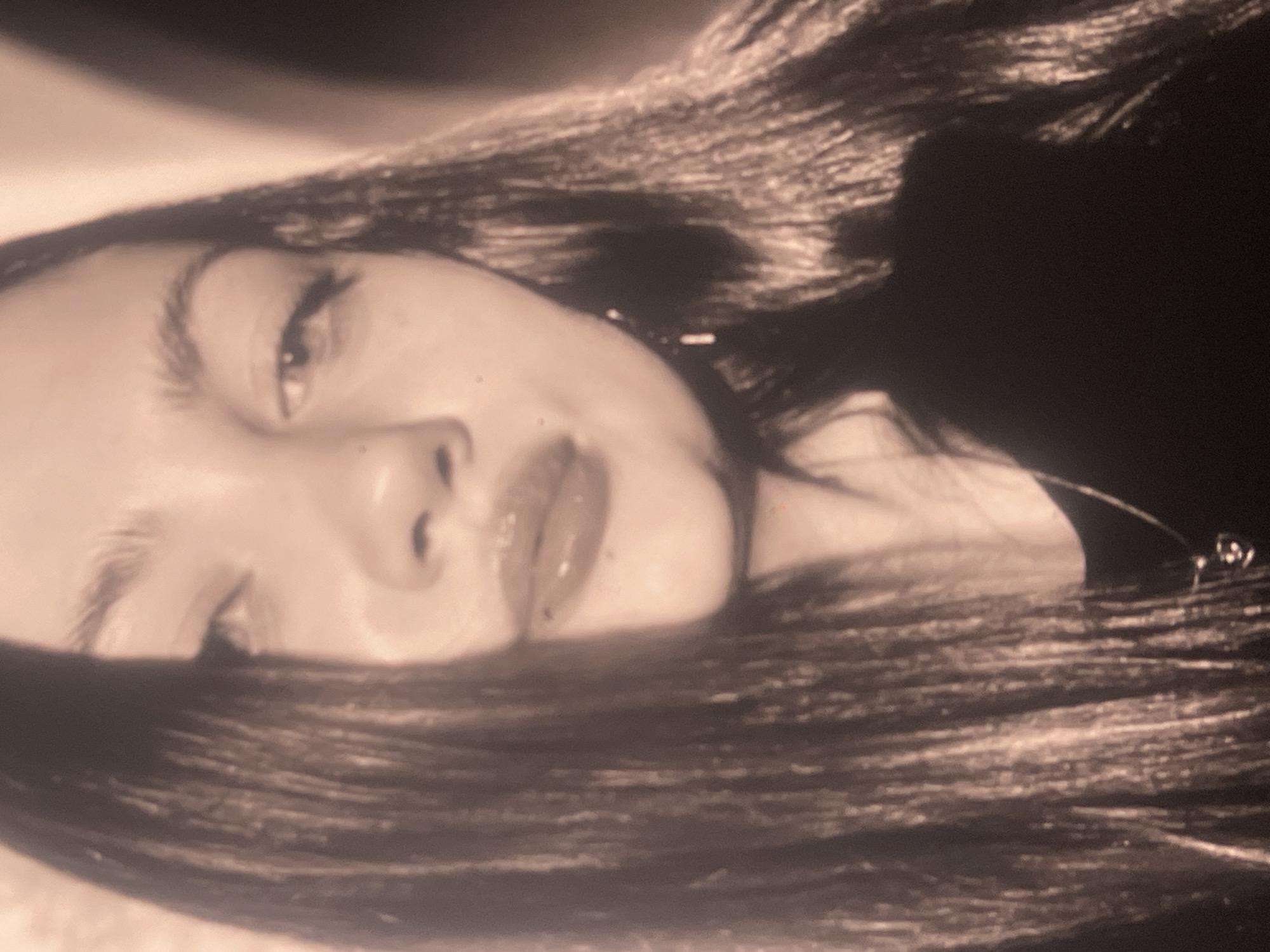 This media file is a close-up, sepia-toned portrait of a person, likely a young woman, with long dark hair. The image is composed diagonally, showing the subject's face from a tilted perspective. The person's face occupies a significant portion of the frame, with abundant dark hair framing the head and cascading around the shoulders.
One eye is partially visible and looking towards the viewer, featuring prominent eyelashes and a defined eyebrow. The other eye is mostly obscured by hair or the angle of the shot. The nose is clearly visible, and the lips are full, appearing slightly parted and glossy. A small mark or mole is noticeable above the upper lip. The skin appears smooth, with some minor texture visible upon closer inspection.
The overall tonality is warm monochromatic, giving the image a vintage or artistic quality. The background is dark and out of focus, emphasizing the subject. The lighting is soft, creating gentle highlights and shadows on the face and hair. The person appears to be posing for the photograph, with a relaxed and direct gaze. No text is visible in the image. There are no specific location cues or time of day discernable from the visual content.