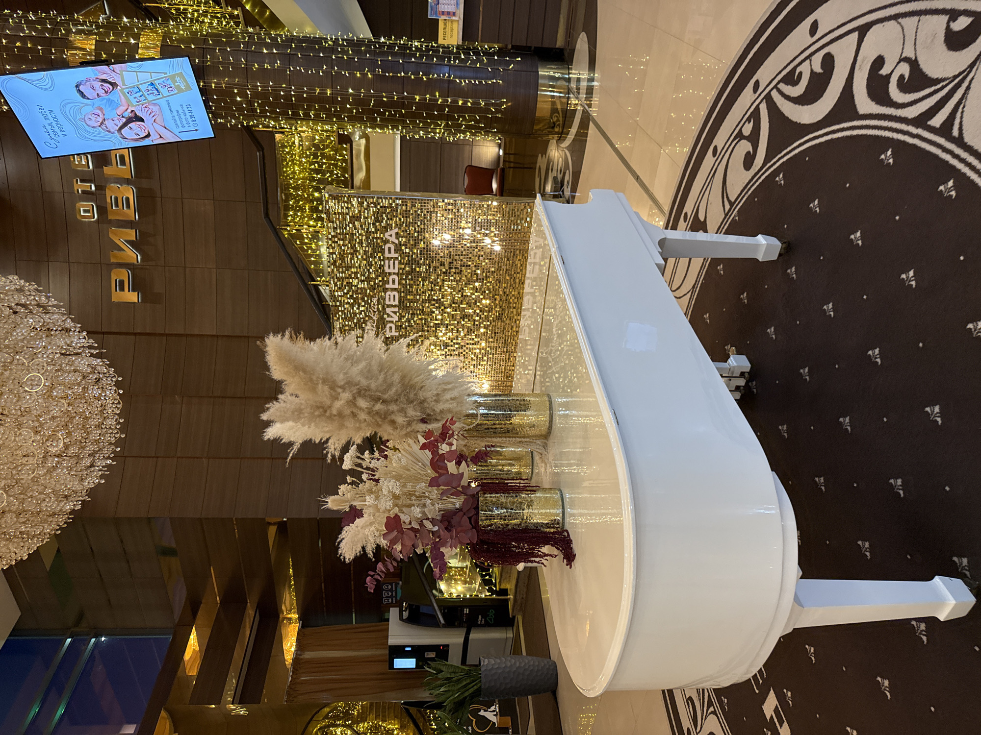 The image depicts the interior lobby of the Riviera Hotel in Kazan, Russia. A white grand piano is positioned on a circular patterned rug in the foreground. Behind the piano, a wall is adorned with a gold sequined backdrop, and further back, a staircase is decorated with strings of golden lights. Above the sequined wall, the hotel's name, "РИВЬЕРА" (Riviera), is displayed in large gold letters. To the right of the hotel name, a digital screen displays an advertisement featuring a family. Various decorative elements, including tall vases with dried reeds and flowers, are placed on the piano. The overall ambiance is one of luxury and festivity, amplified by the warm lighting and shimmering decor.