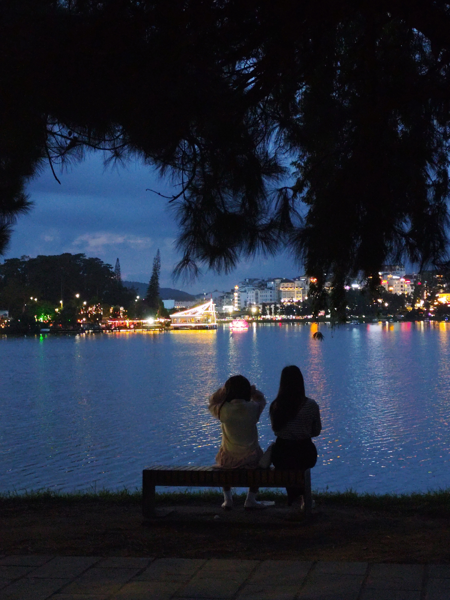 Two females sit on a bench overlooking Ho Xuan Huong Lake in Da Lat, Vietnam, at night. The city lights reflect on the water's surface. Dark branches frame the upper portion of the image.