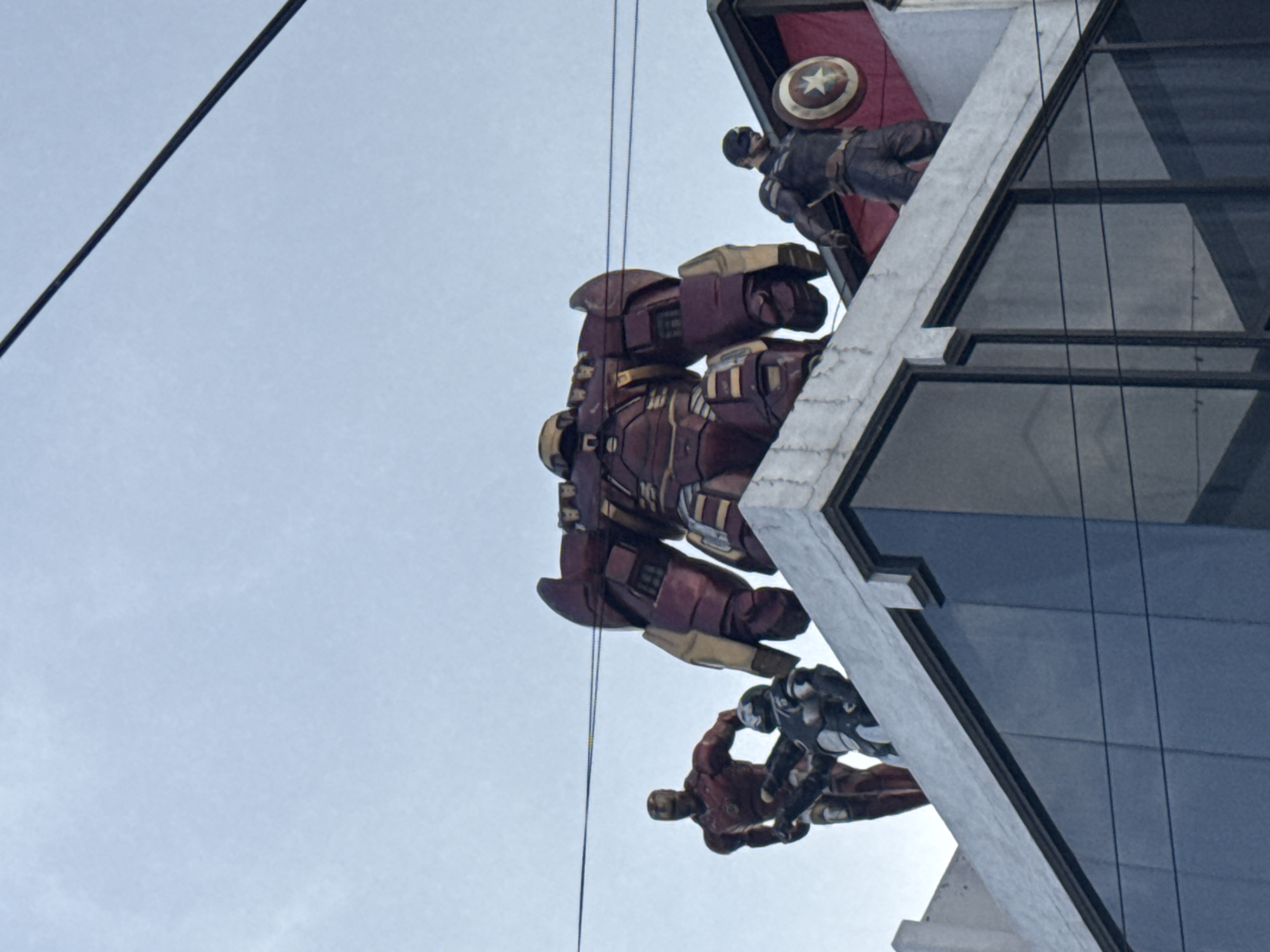 The image, taken from a low angle, features a building in Mexico City, Mexico, adorned with large-scale figures of Marvel superheroes. At the top of the building, a statue of Captain America holding his shield is visible, set against a red and dark-red backdrop. Below, a substantial Hulkbuster figure is mounted. Near the base of the visible section of the building, there are figures of Iron Man and War Machine. The building itself appears to be of modern design, featuring glass windows and light gray concrete architectural elements. Electrical wires cross the view, cutting across the sky. The sky is a pale blue.
