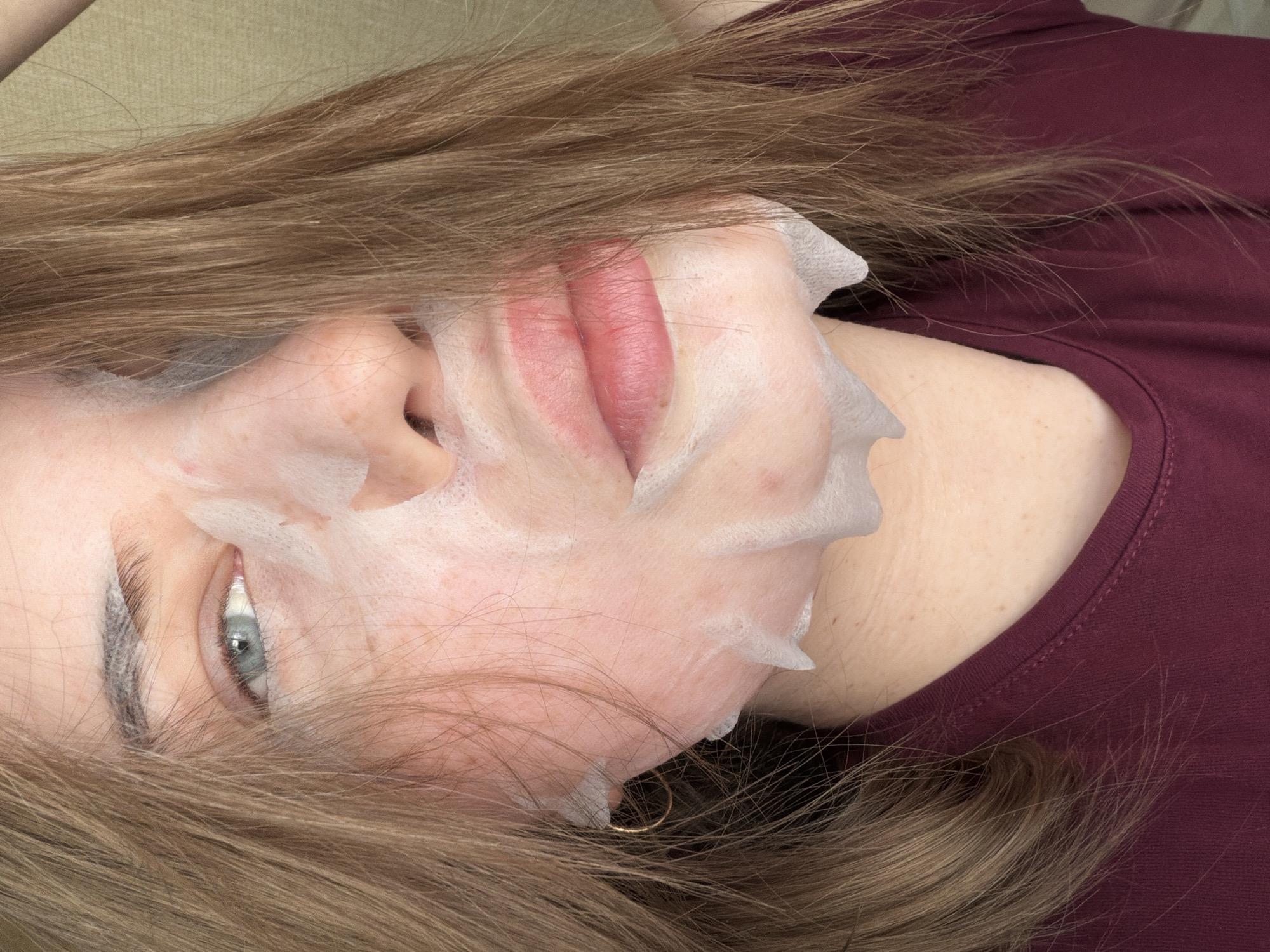 A light-skinned person with blonde hair is lying on their back. They have a white sheet mask on their face, covering the nose and mouth area. Their left eye is open and blue. The person is wearing a maroon-colored t-shirt. A gold earring is visible near the ear. The background is a neutral beige color. No features suggest the location is Chaikovsky, Russia.