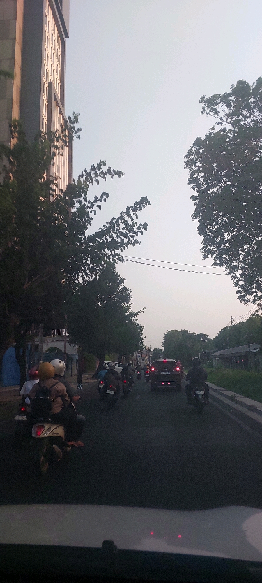 The image depicts a street scene in Surabaya, Indonesia, characterized by a moderate traffic flow of motorcycles and cars. On the left side of the road, a tall, modern building with a grid-like facade is visible, partially obscured by the branches of lush green trees. The trees line the street, creating a canopy effect in some areas.
The road itself is asphalt and appears to be a multi-lane street. Several motorcycles are in motion, with riders wearing helmets in various colors, including white, yellow, and red. Some motorcycles are carrying passengers. Ahead, a dark-colored SUV is stopped in traffic, its brake lights illuminated. Other vehicles, including more motorcycles and cars, are visible further down the road, suggesting a typical urban traffic condition. The sky is overcast with a pale, hazy light, indicative of either early morning, late afternoon, or a hazy day. The scene is captured from the perspective of someone inside a vehicle, with the dashboard or hood visible in the foreground, out of focus. The overall atmosphere is that of a busy urban environment during daylight hours.