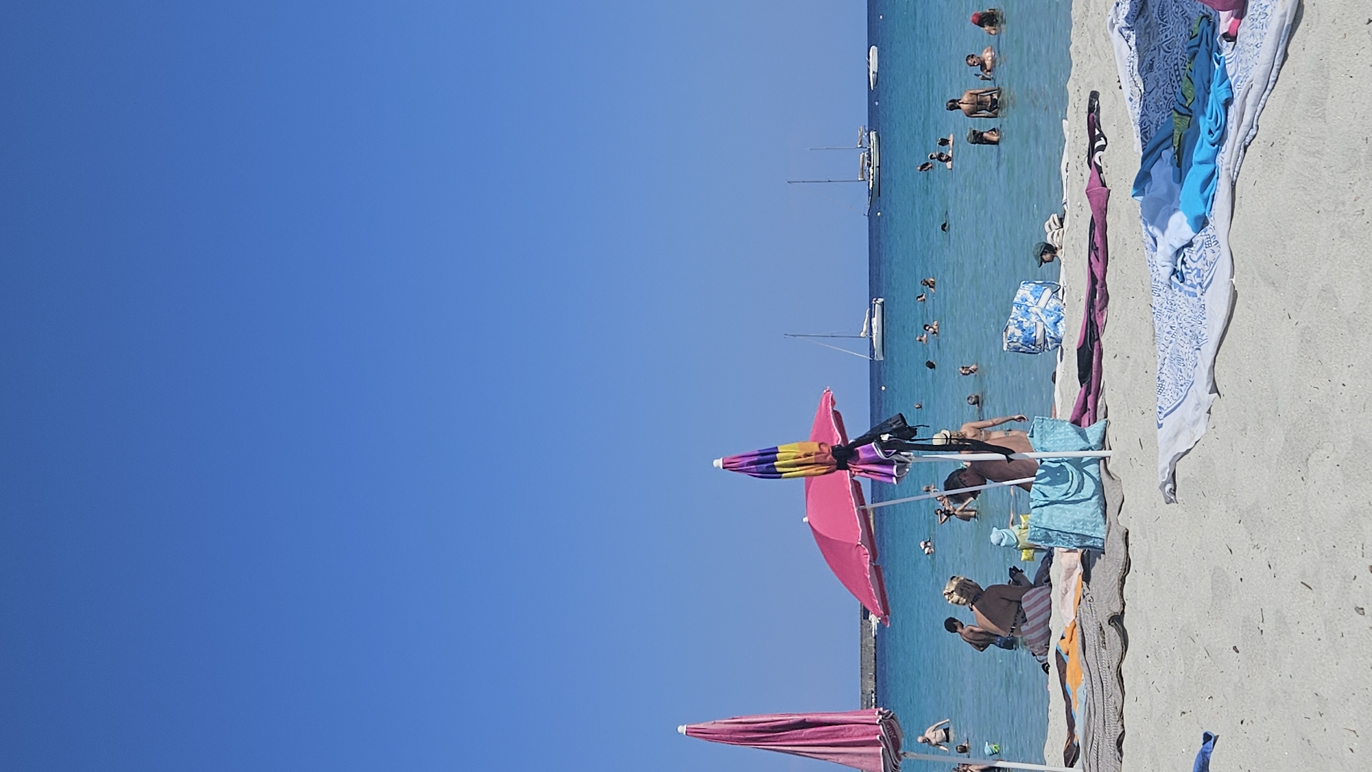 The image captures a sunny day at a beach, likely in L'Île-Rousse, France, given the clear blue sky and turquoise waters. In the foreground, several beach towels and bags are spread on the sandy shore. Two vibrant pink beach umbrellas are prominent, one fully open and the other partially obscured.
Further back, the shallow, clear water teems with activity. Numerous people are visible swimming, wading, and enjoying the sea. A few individuals are seated on the sand near the water's edge. In the middle ground and background, several sailboats are anchored or cruising on the calm, blue ocean. A long, low structure, possibly a pier or a breakwater, stretches across the horizon on the left side of the image. The overall scene depicts a leisurely and lively beach day.