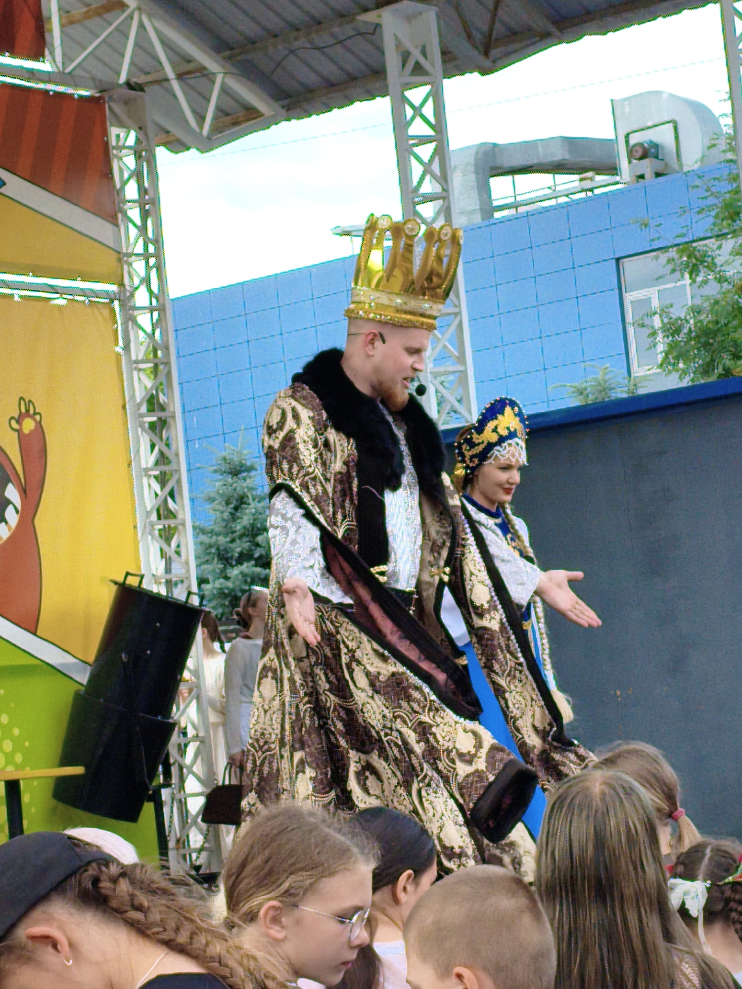 The image depicts a festive outdoor event, likely a performance or parade, with people in costumes on a stage. A man wearing a golden crown and an ornate, fur-trimmed robe stands prominently, speaking into a microphone. Beside him, a woman in a traditional blue and gold headdress and a similarly decorated garment gestures with her hand. Behind them, a blue building with a grid pattern is visible, along with trees and scaffolding. In the foreground, a crowd of spectators, including children, are gathered, some looking towards the stage, while others are focused on something off-camera. The overall impression is one of public celebration and entertainment. The location is the city of Orenburg, Russia.