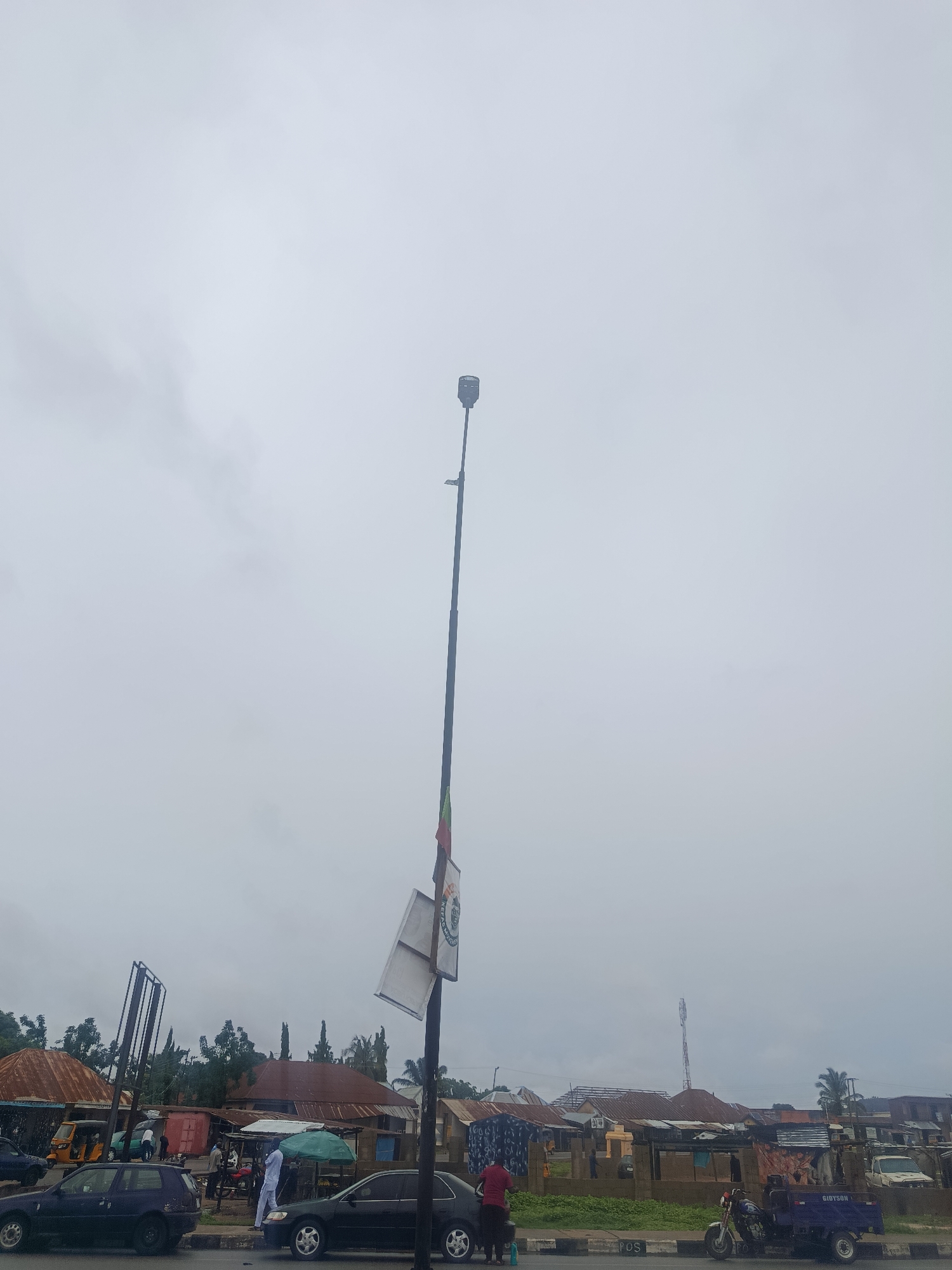 The image depicts a cloudy day in Jalingo, Nigeria, likely on a street with a few shops and a marketplace. A tall street lamp stands prominently in the foreground, adorned with a flag and two white signs, one of which is angled. Several vehicles are present, including a dark blue car, a black sedan, a yellow tricycle, and a blue tricycle with the word "GIBSON" visible on its side. People are seen near some of the stalls, some appearing to be vendors or shoppers. The background shows buildings with corrugated iron roofs, typical of the region, and lush green trees. The overall atmosphere suggests a typical day in a Nigerian town.