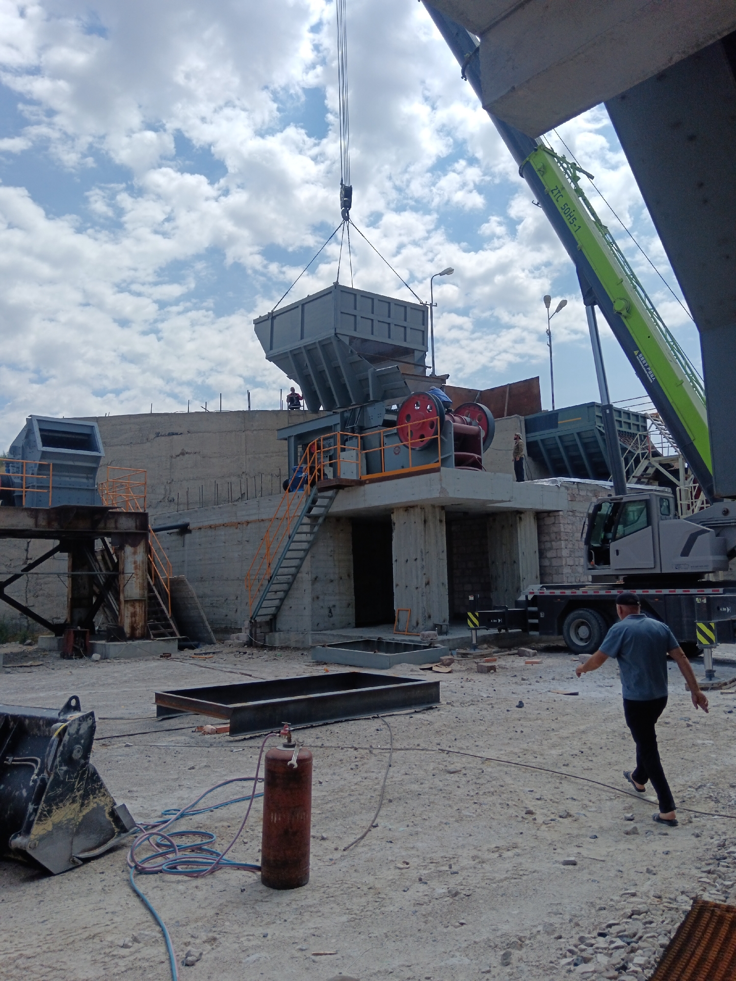 The image depicts a construction site, possibly in Aragatsavan, Armenia, under a cloudy sky. In the foreground, a person in a blue t-shirt and black pants walks away from the camera. To the left, a large black excavator bucket lies on the ground, next to a red oxygen tank with hoses attached. In the mid-ground, industrial machinery, including a large crusher with red wheels, is visible. A crane with a green boom extends into the frame from the right, supporting a gray bin that is being lowered by another crane in the background. Several metal structures and scaffolding are also present, indicating ongoing construction work. The ground is covered with dirt, gravel, and debris.