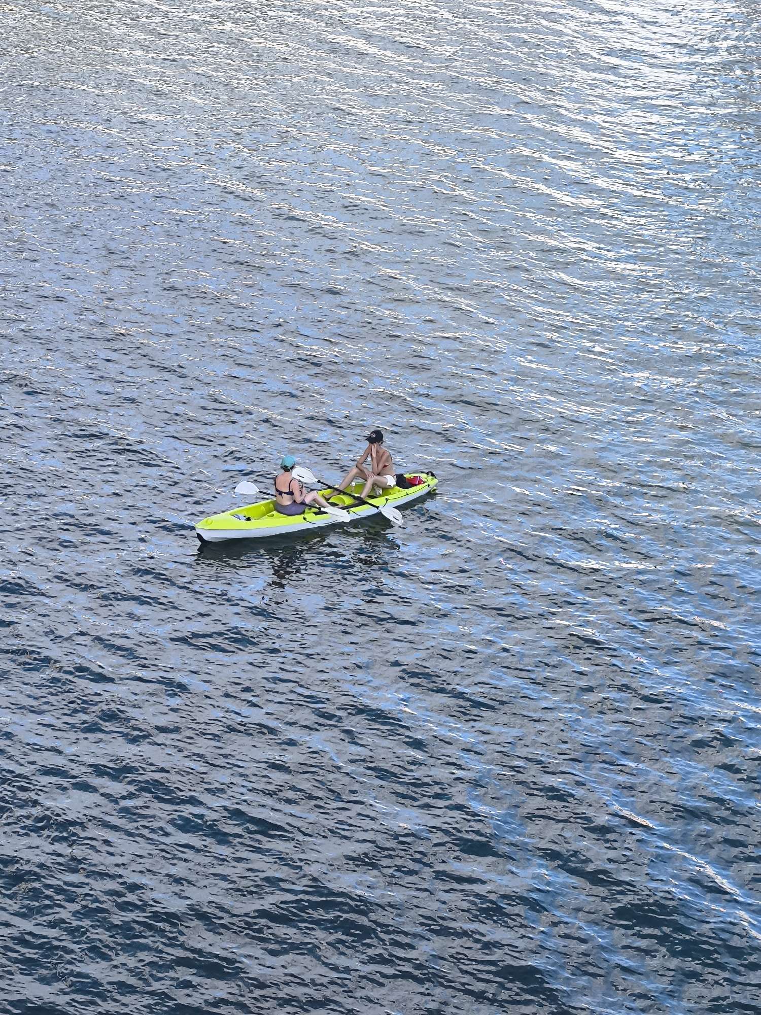 The image shows two people in a yellow kayak on a body of water. One person is in the front, wearing a blue bikini top and a blue head covering, and is holding a paddle. The other person is in the back, wearing a red bikini and a dark baseball cap, and is also holding a paddle. The water is a deep blue with light reflecting off the surface, creating ripples. The kayak is positioned diagonally in the frame, with the bow pointing towards the upper left. There is no indication of the location being in Vienna, Austria.
