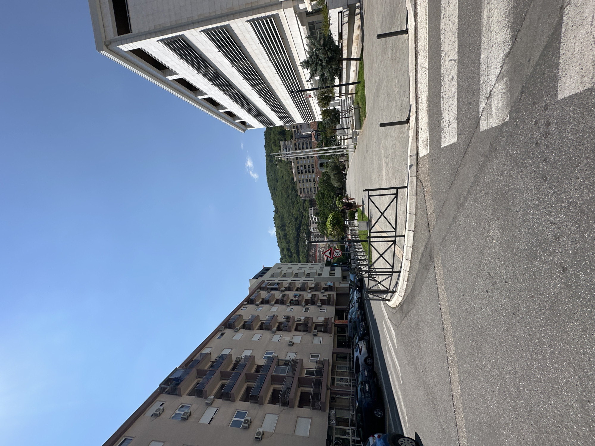 The image shows an outdoor scene in Budva, Montenegro, likely during the daytime due to the bright sunlight and clear blue sky. The perspective is from a low angle, looking up and sideways. In the foreground, a paved road with a pedestrian crossing is visible. To the left of the road, a dark metal fence with a diamond pattern separates the sidewalk from the street.
Dominating the left side of the frame is a modern, multi-story building with a light-colored facade and horizontal windows. The building's architecture suggests an office or residential complex. Further back, across the street and beyond a line of trees, another, older, mid-rise apartment building is visible. This building has numerous balconies with dark railings. Behind these buildings, a lush green, tree-covered mountain slopes upwards, indicating a hilly terrain.
Along the side of the road, several cars are parked, mostly dark-colored. A few people are walking on the sidewalk in the distance. A traffic sign with "30" inside a red circle is visible, indicating a speed limit of 30 kilometers per hour. The overall impression is of a well-maintained urban environment integrated with natural scenery. The weather appears to be clear and sunny.