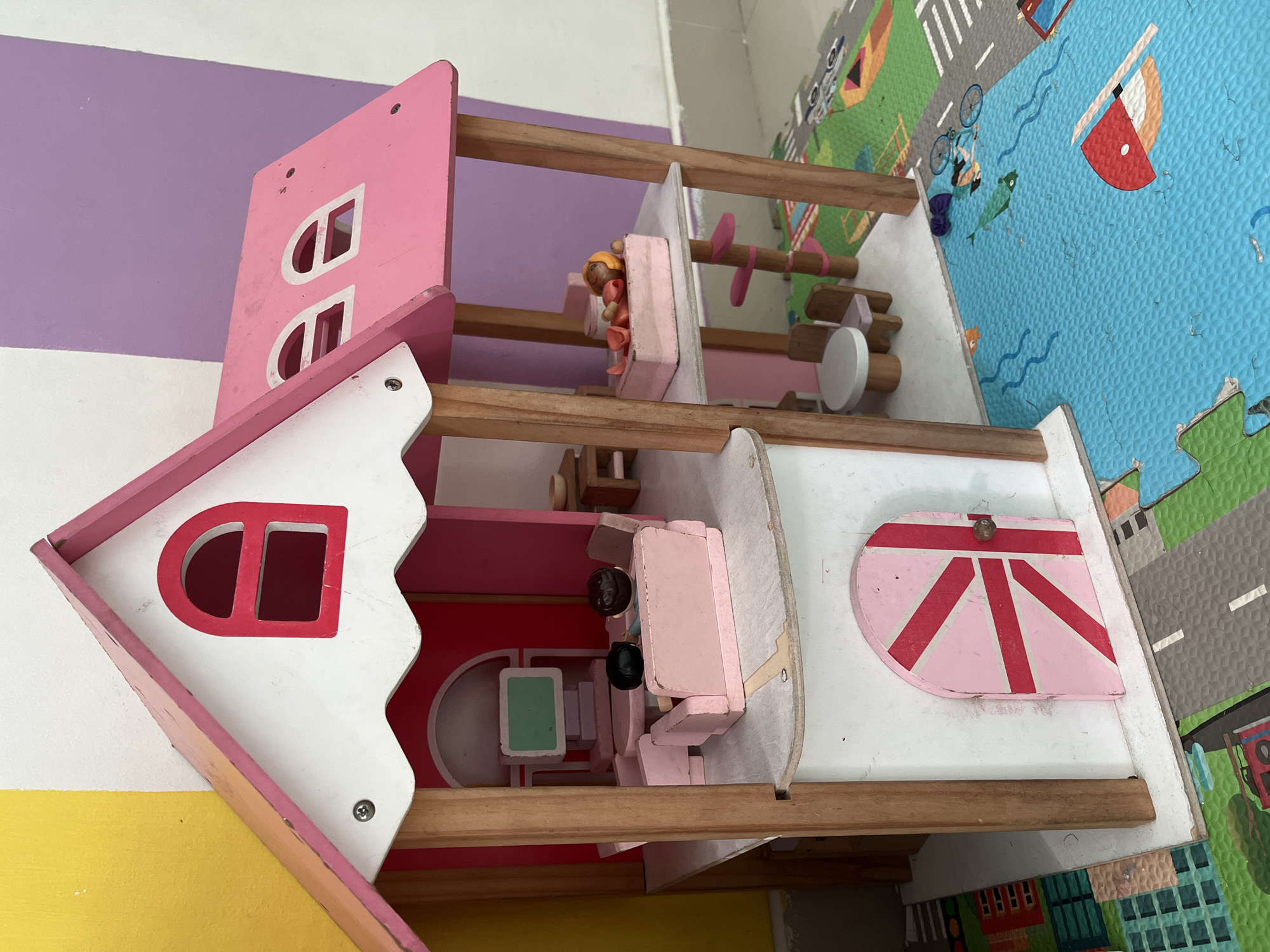 This is a close-up, eye-level, full shot of a pink and white wooden dollhouse situated on a colorful play mat. The dollhouse has two stories, with a pink roof and white walls. It features several windows and a pink door with a red Union Jack design. Inside the dollhouse, miniature furniture and two small dolls are visible. The dollhouse is positioned on a blue mat depicting a body of water with boats and fish, and green mats depicting a cityscape with roads and buildings. The background consists of a wall with vertical stripes of yellow, white, and purple. The lighting is bright and even, suggesting an indoor setting. There is no discernible information in the image that would indicate the location in the city of Kuta, Indonesia.