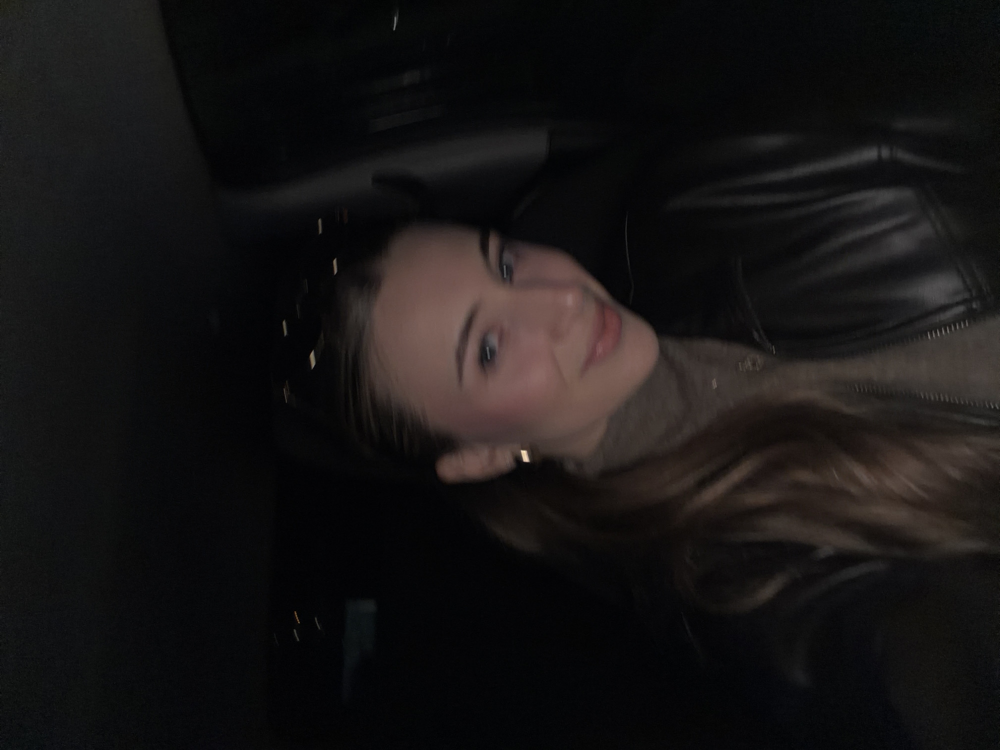 A young adult female is depicted in a close-up, front-facing portrait taken inside a vehicle at night. She has light skin, blue eyes, and light brown hair styled into a ponytail. She wears a brown turtleneck top under a black, possibly leather, jacket. A small gold-colored hoop earring is visible on her right ear. Her expression features a subtle smile, with light pink blush visible on her cheeks and a soft lip color. The primary light source illuminates her face from the front. The rest of the vehicle's interior and the background are largely obscured by darkness. The background shows indistinct, blurred pinpricks and streaks of light, consistent with distant city lights or passing vehicle lights at night. The environment is the interior of a vehicle during nighttime. No specific landmarks or geographic features are visible within the image to establish a precise location within Plovdiv, Bulgaria.